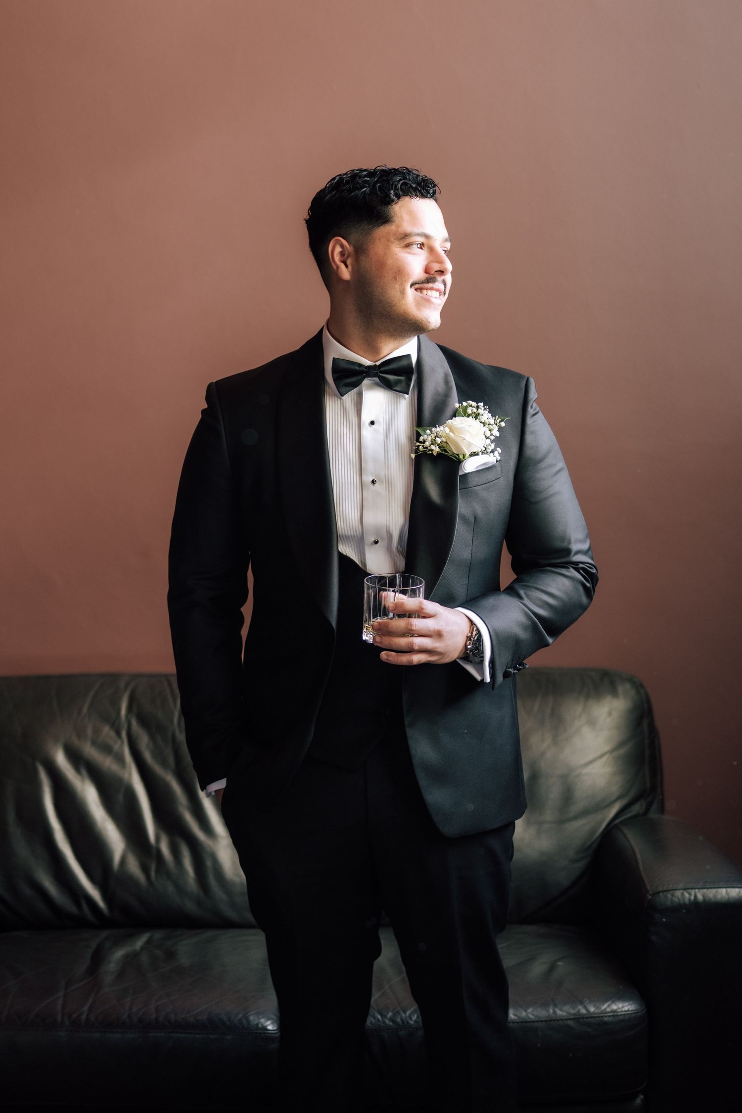 Smiling groom in black tuxedo with boutonniere holding a glass while leaning against a leather sofa.