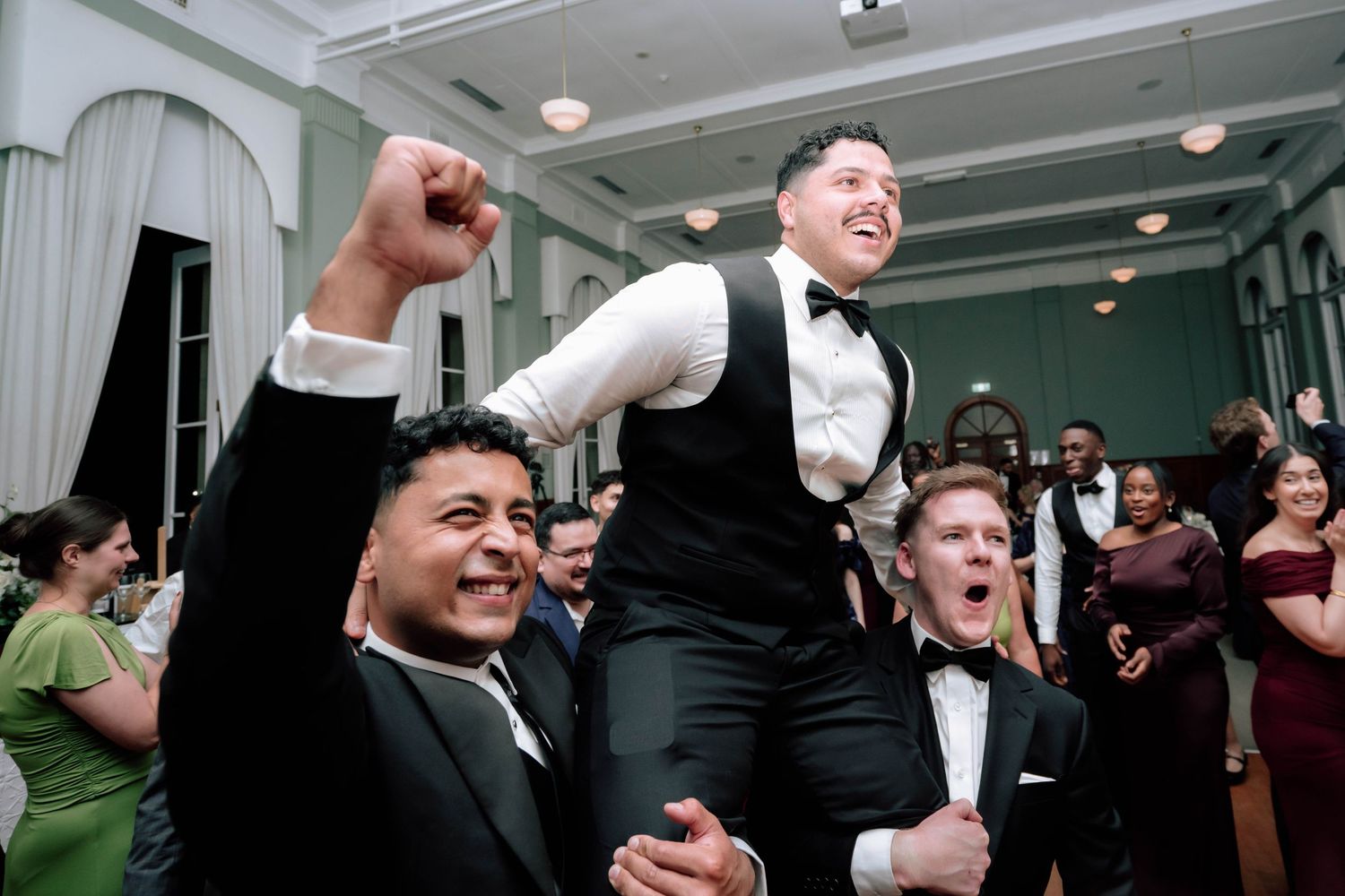 Groom lifted on shoulders by groomsmen celebrating on wedding dance floor.