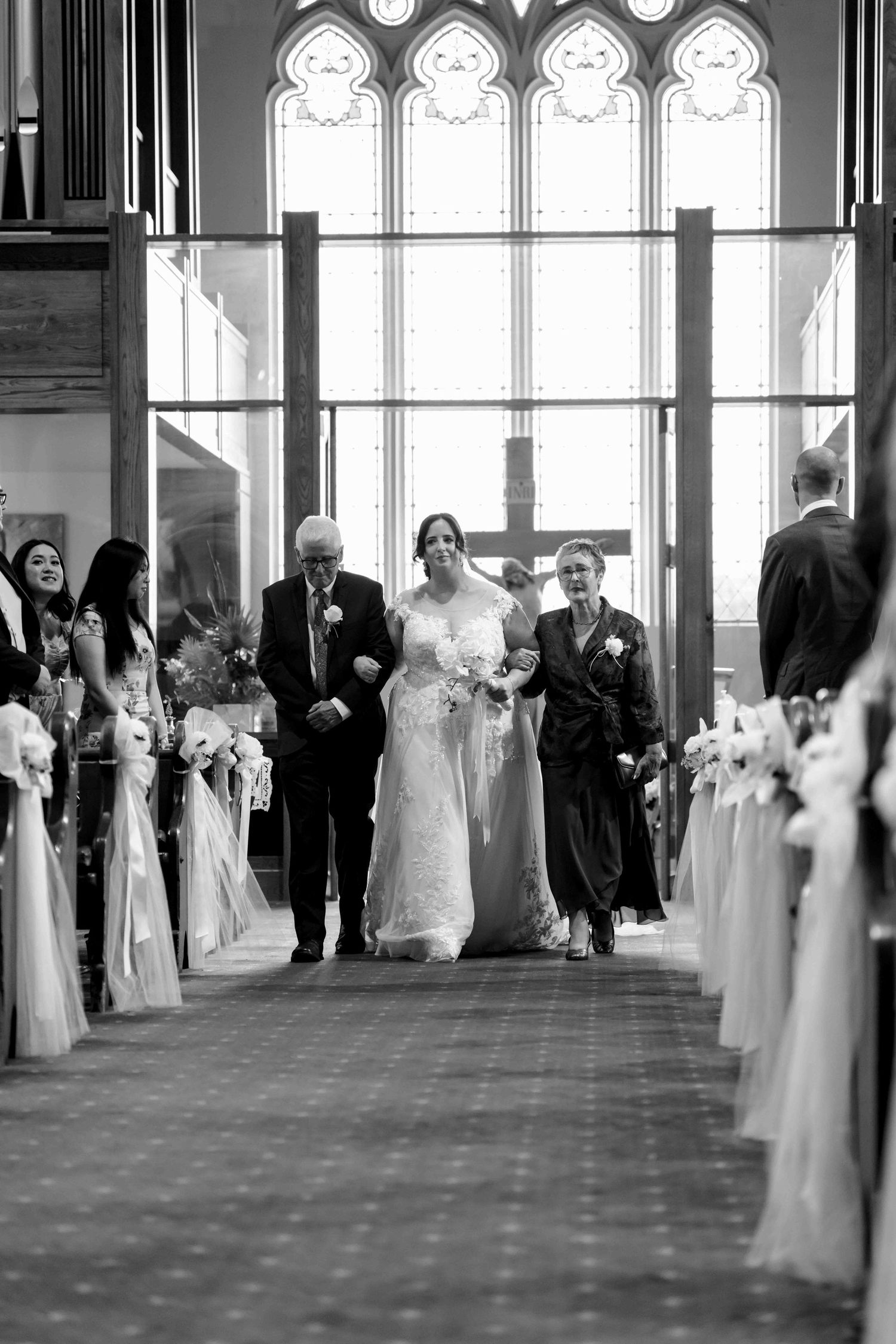Bride walks down church aisle escorted by two family members in black and white ceremony photo.