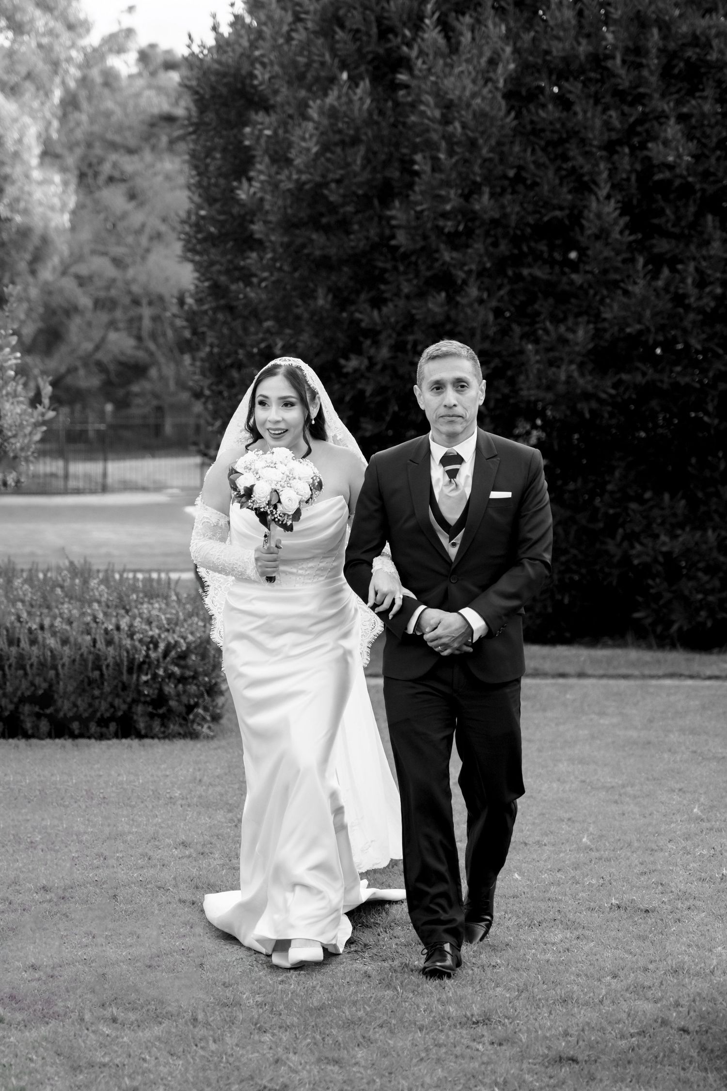 Bride and groom walk together across lush garden lawn in timeless black and white wedding portrait.