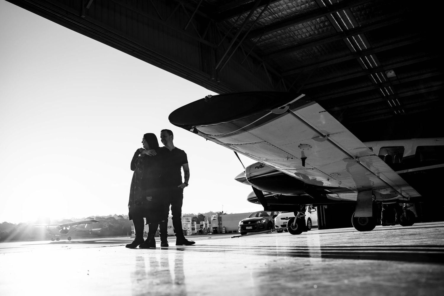Black and white silhouette of a couple embracing in an airplane hangar with bright sunlight behind them.