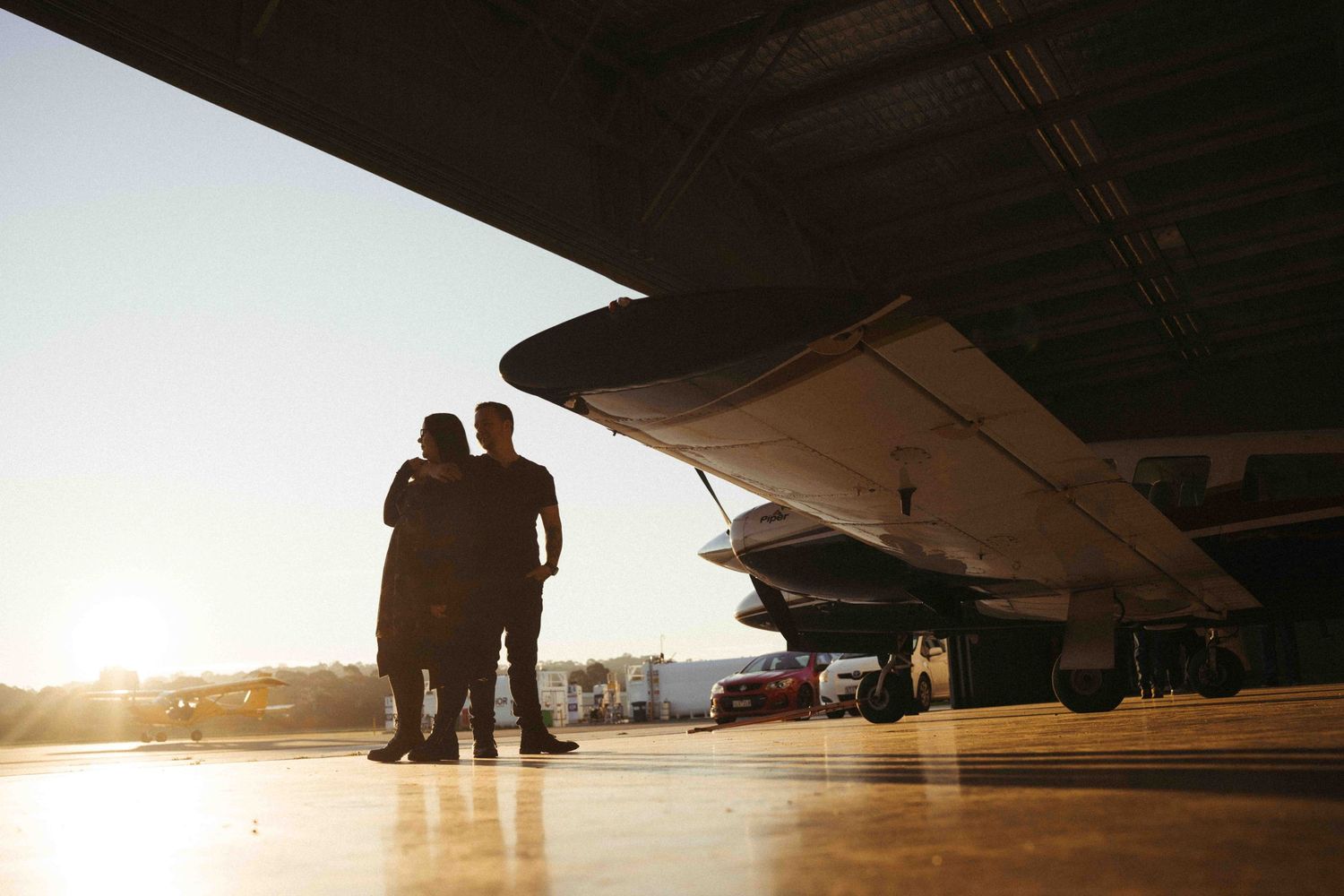 Two silhouetted figures stand beneath a large aircraft wing at sunset on an airport tarmac.