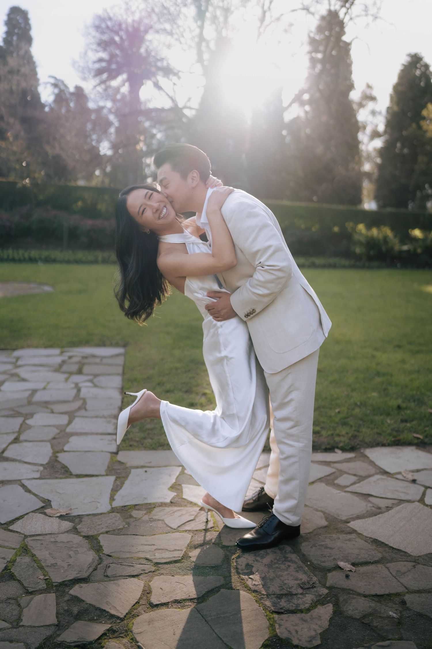 Couple in white wedding attire embracing on stone path in sunlit garden, groom kissing bride as she lifts her foot.