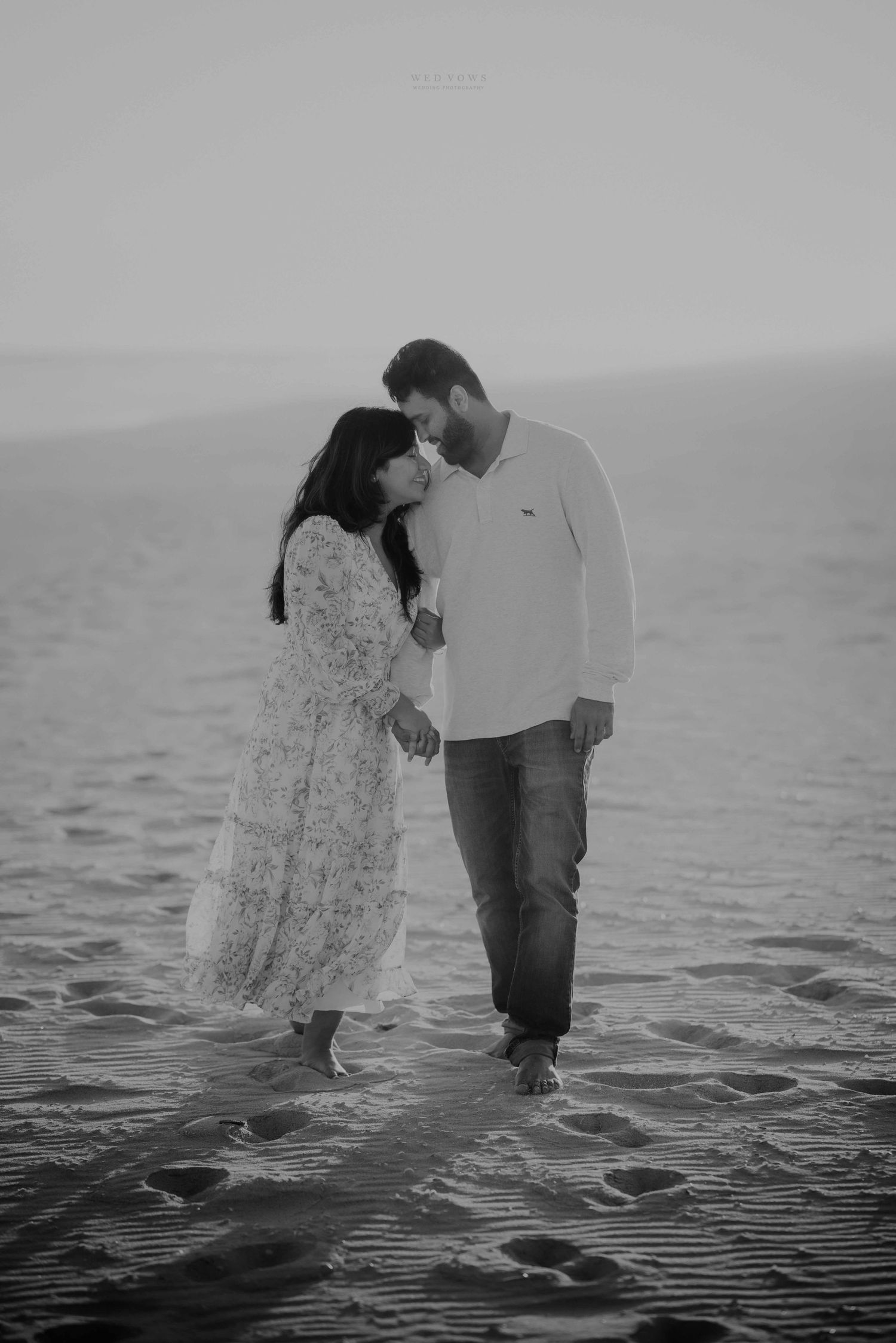 Black and white photo of a couple tenderly touching foreheads while holding hands on a beach.