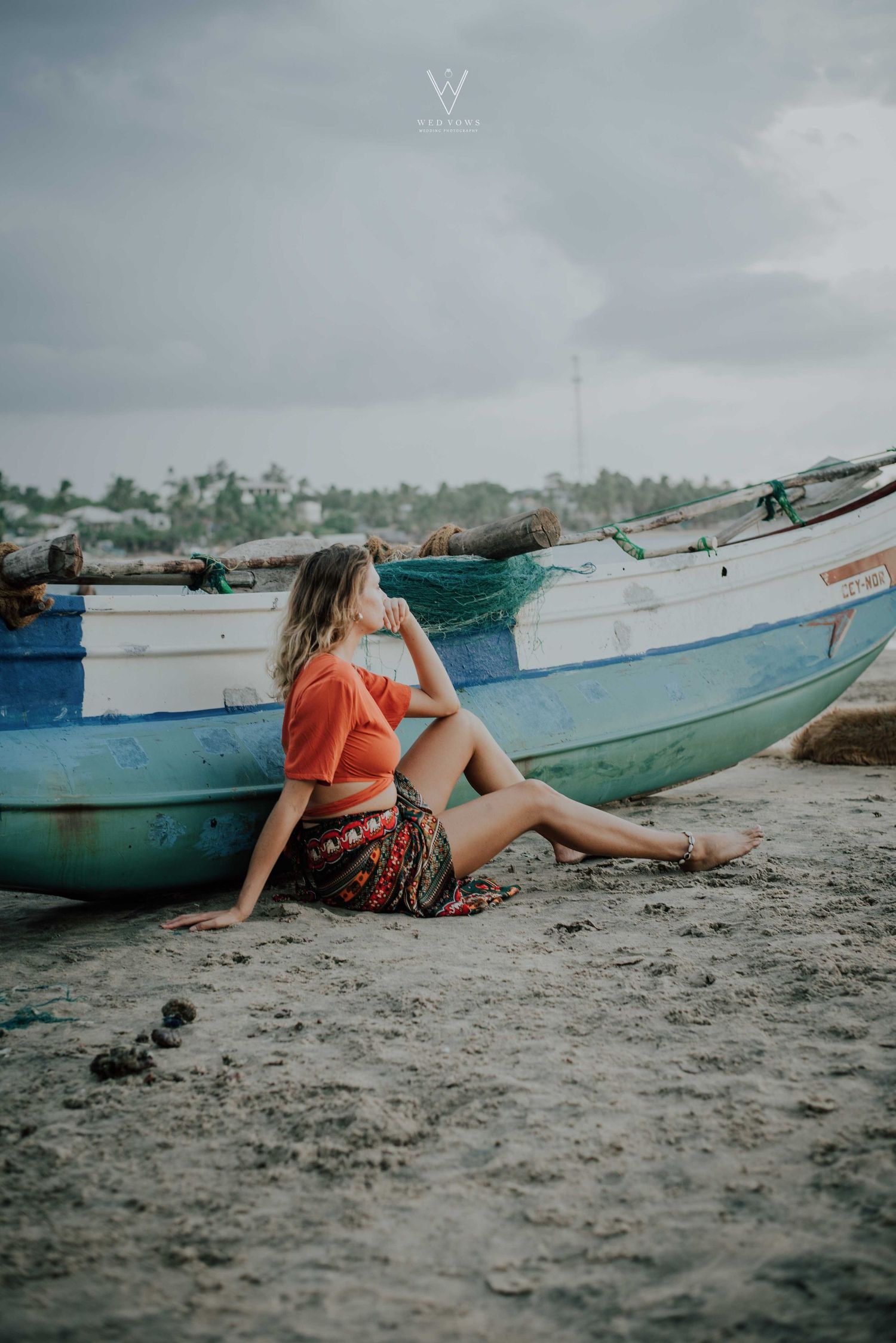 Woman in orange crop top sitting on sandy beach leaning against a colorful blue fishing boat.