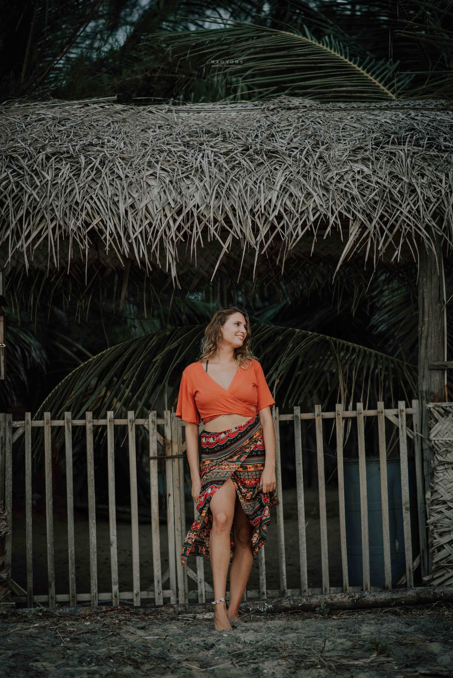 A woman in an orange top and colorful skirt stands before a rustic thatched bamboo fence amid tropical palms.