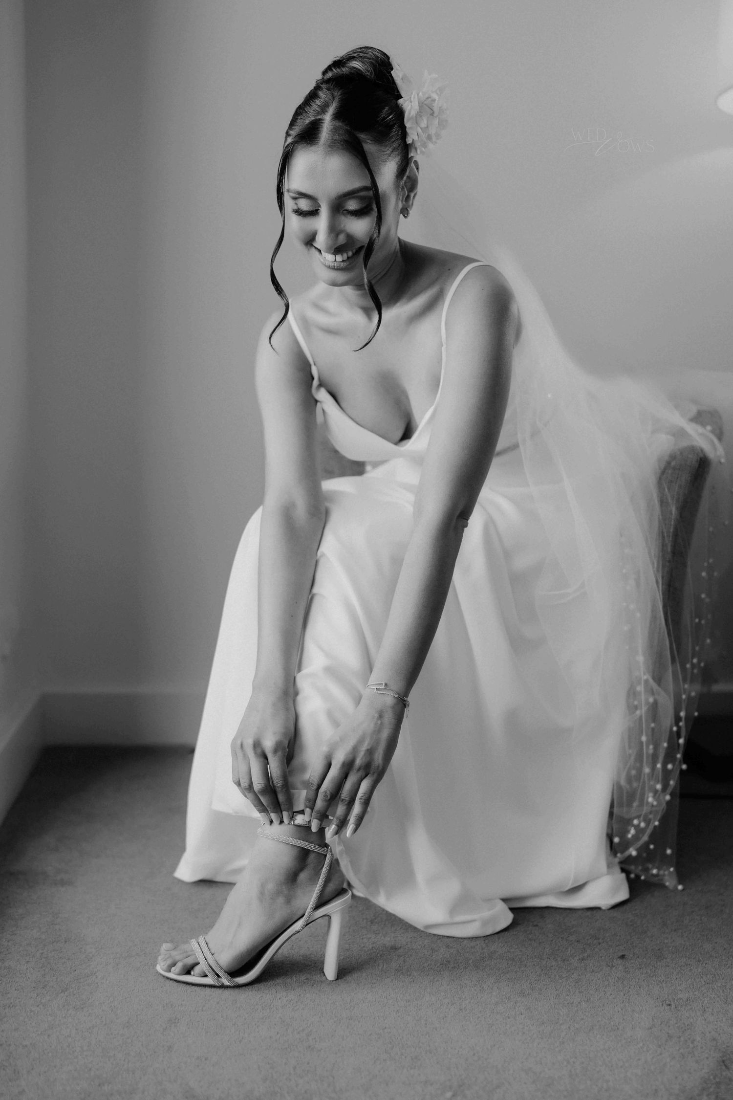 Smiling bride in white gown adjusting her strappy heels while seated, wearing a veil and hair flower.