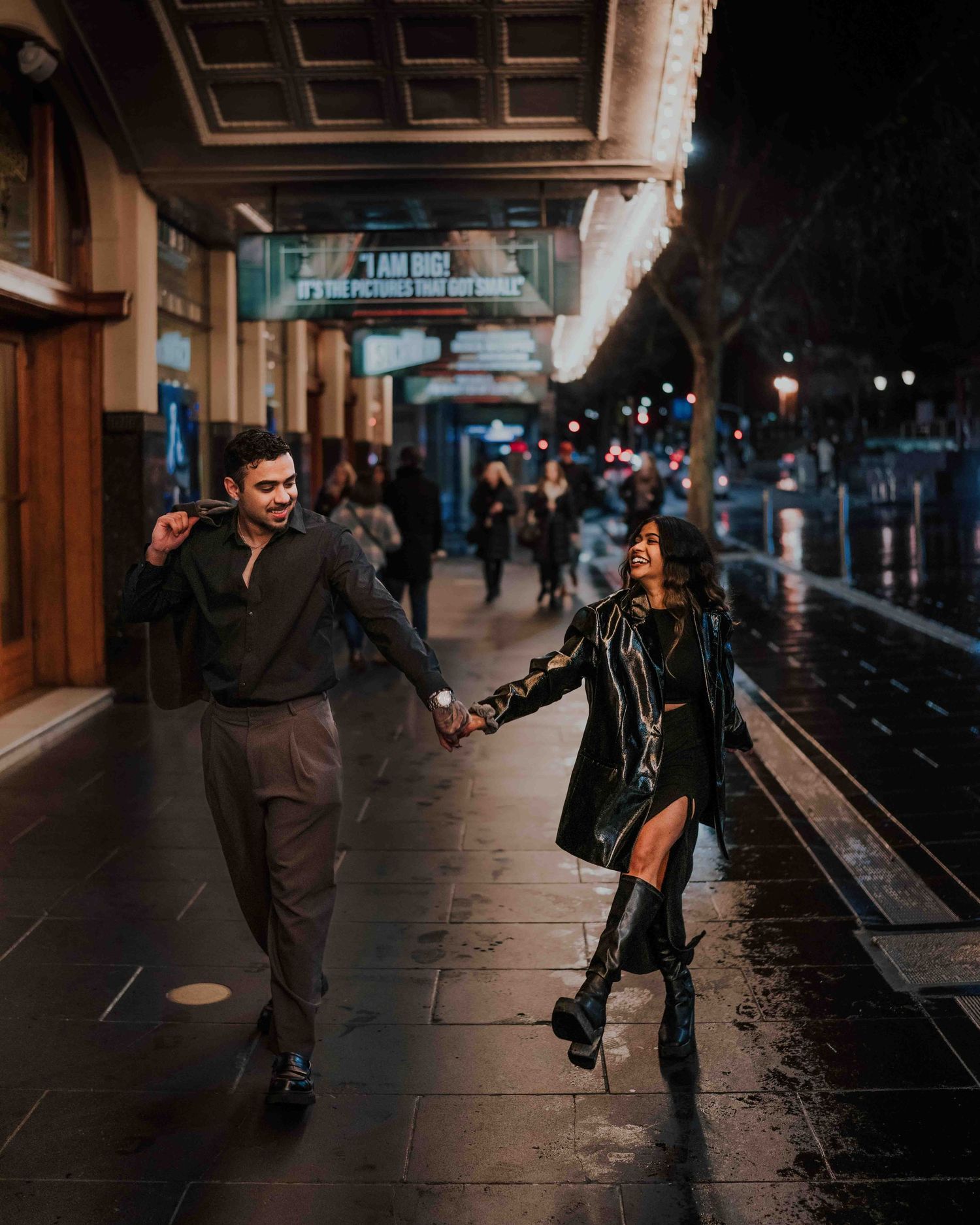 Couple dancing and holding hands on a rainy night street outside a lit theater marquee.