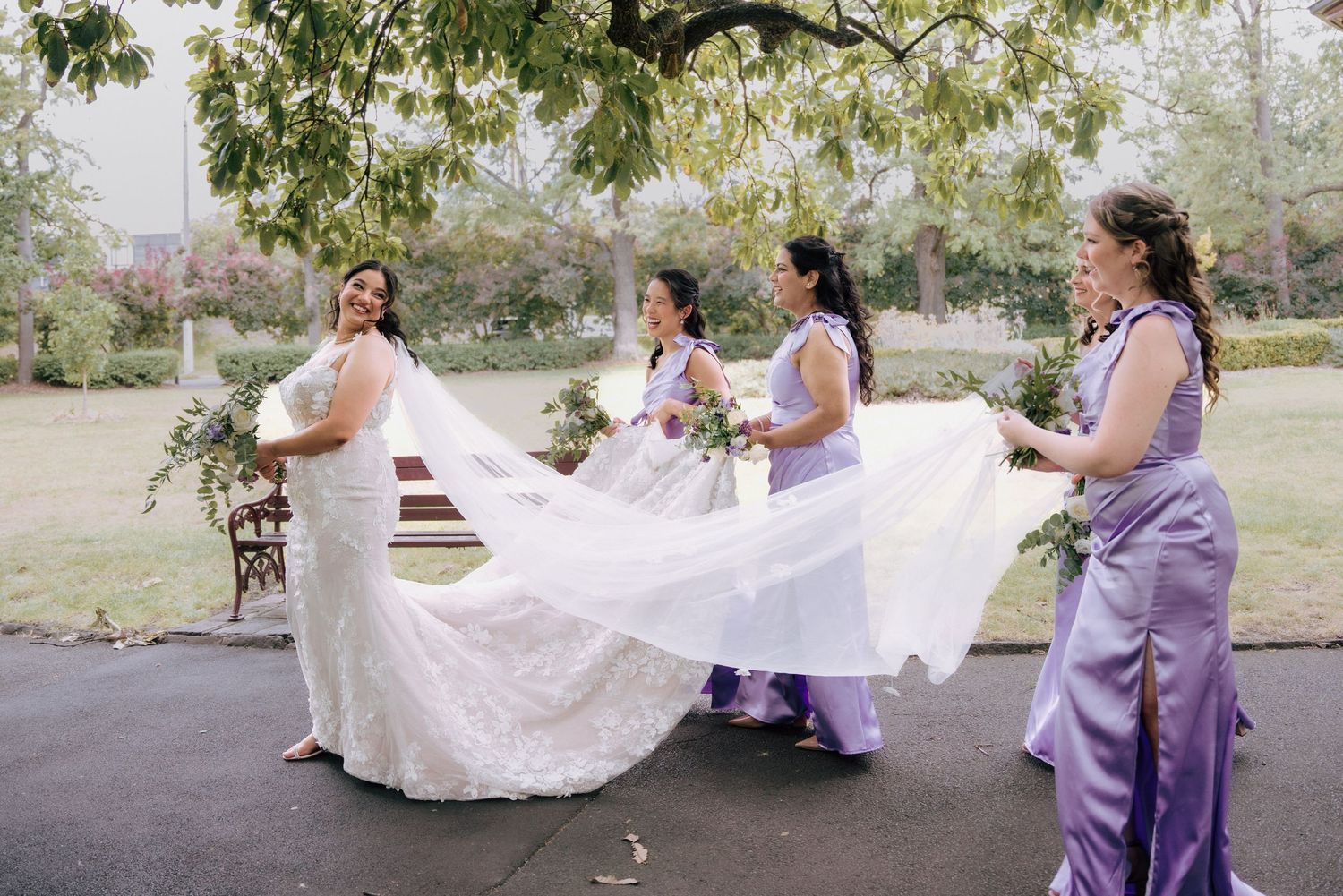 Bride in lace gown walks with three bridesmaids in lavender dresses carrying her long flowing veil.