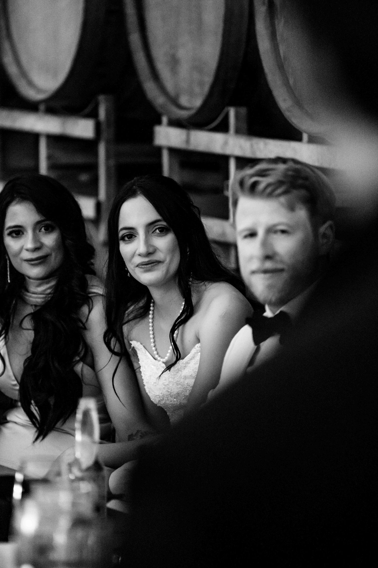 Black and white photo of bride in pearl necklace seated at wedding reception table with guests.