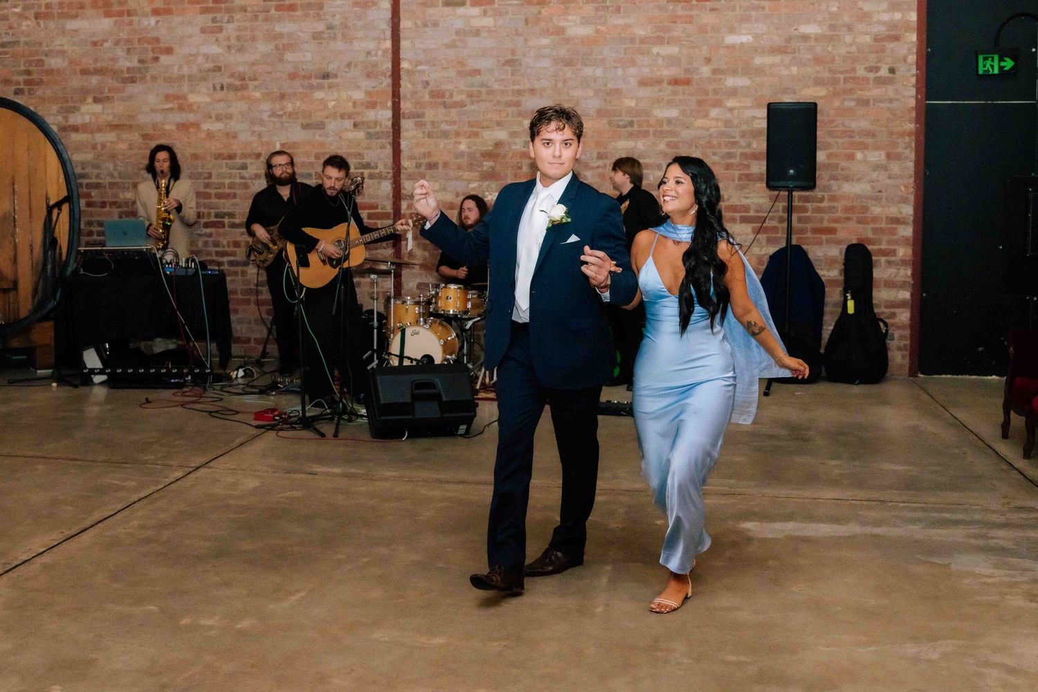 Groom in navy suit dances with bridesmaid in blue dress while live band performs at wedding reception.