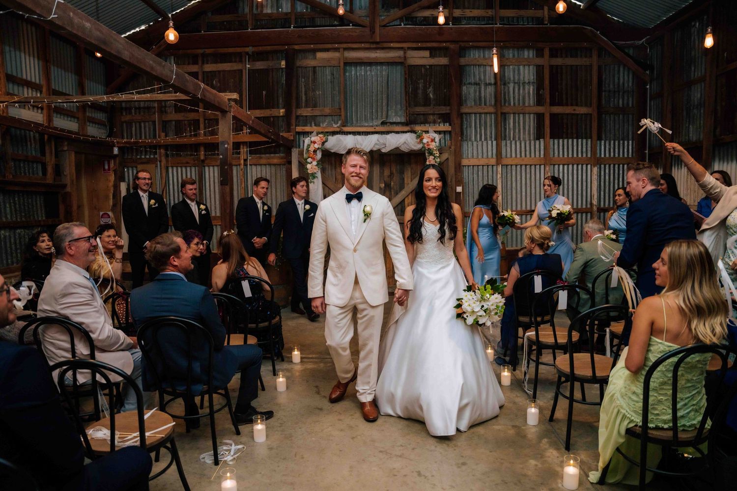 Newlyweds walk hand in hand down the aisle in a rustic barn wedding venue surrounded by seated guests.