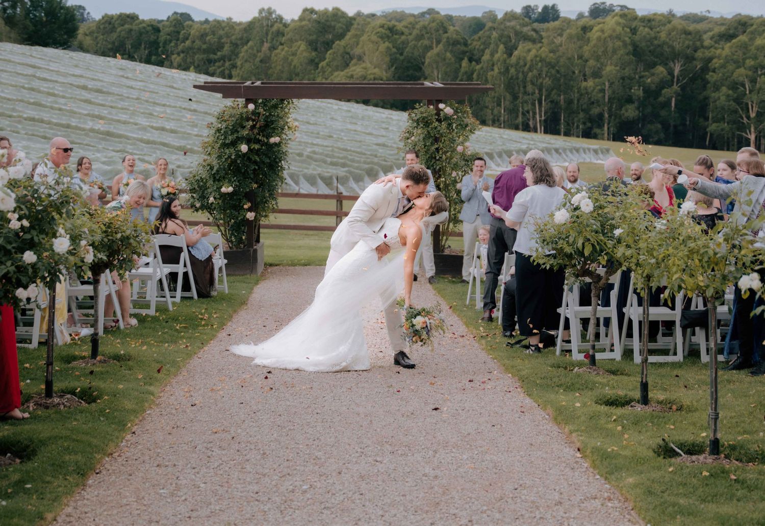 Newlyweds share a dip kiss at outdoor garden wedding ceremony aisle.