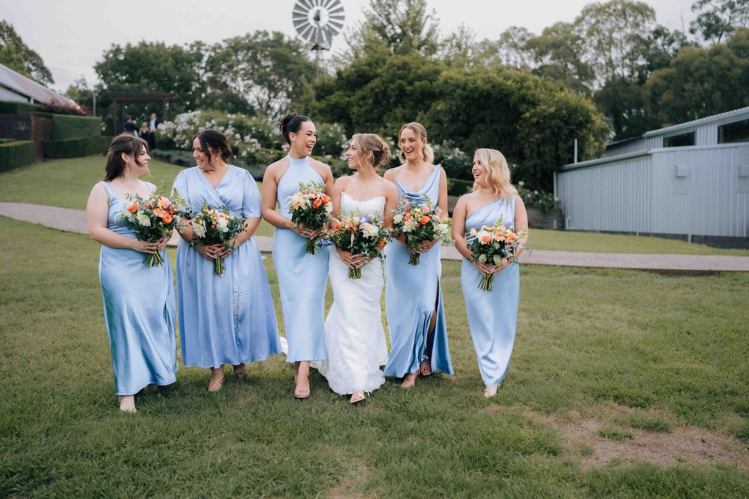 Bride and five bridesmaids in blue satin gowns walking together on grass with windmill backdrop.