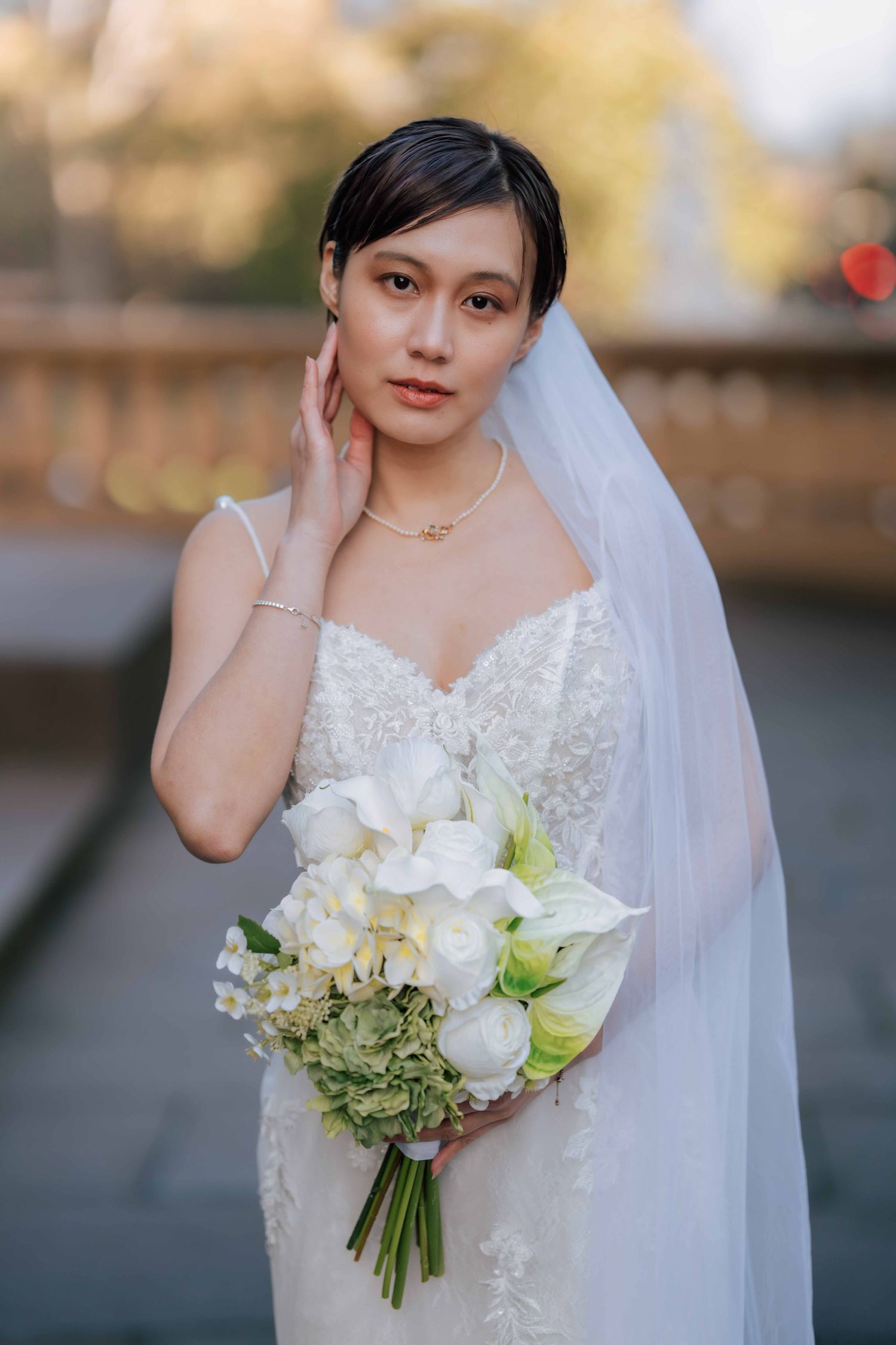 Beautiful Asian bride in lace gown and white veil holds white and green bouquet, posing gracefully outdoors.