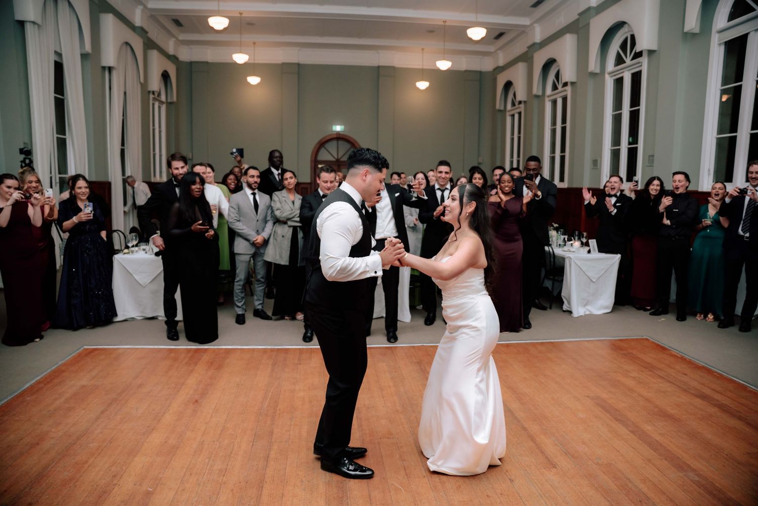 Newlyweds share first dance on wooden dance floor surrounded by applauding wedding reception guests.