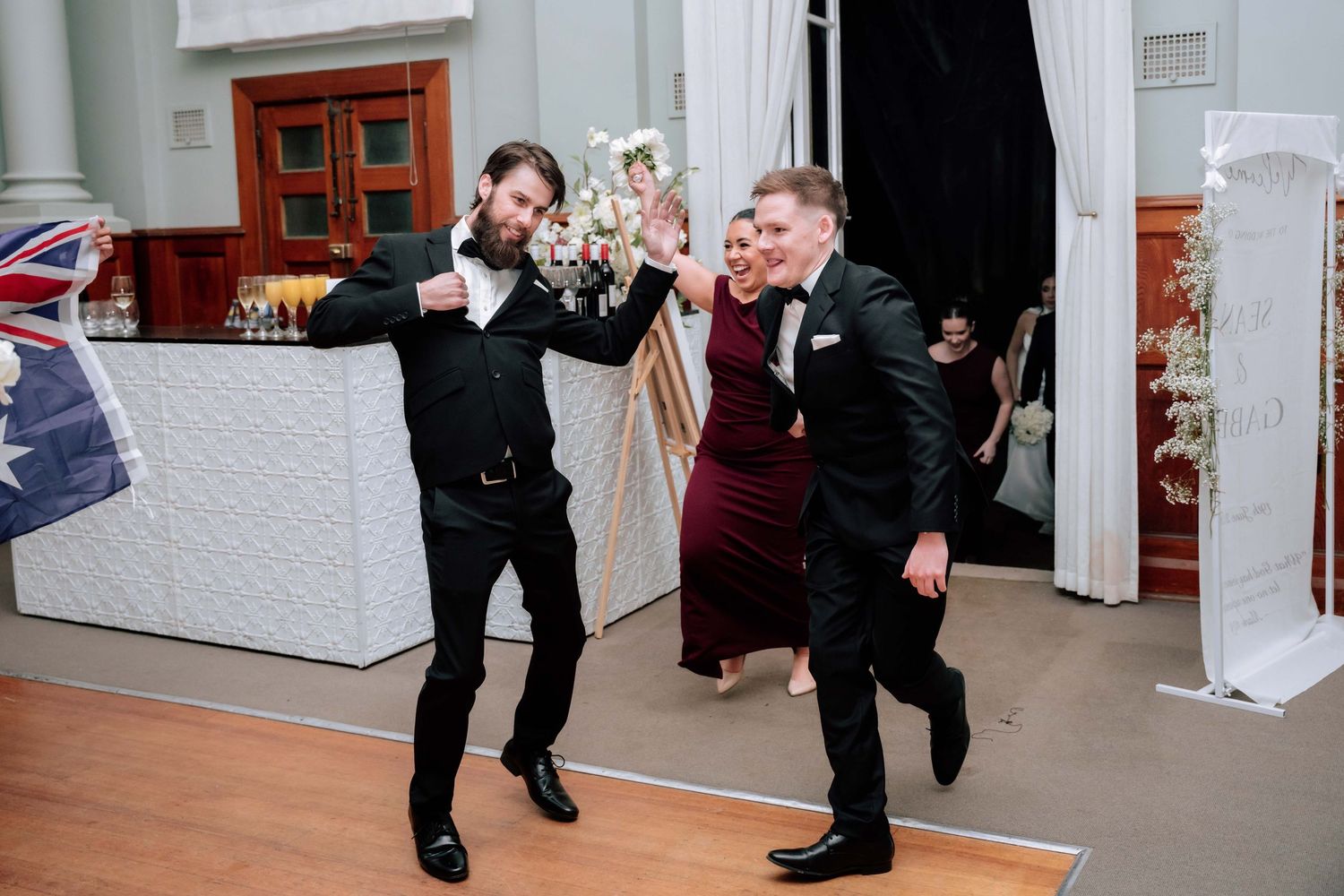 Two groomsmen in black suits celebrating energetically at wedding reception near floral arrangement.