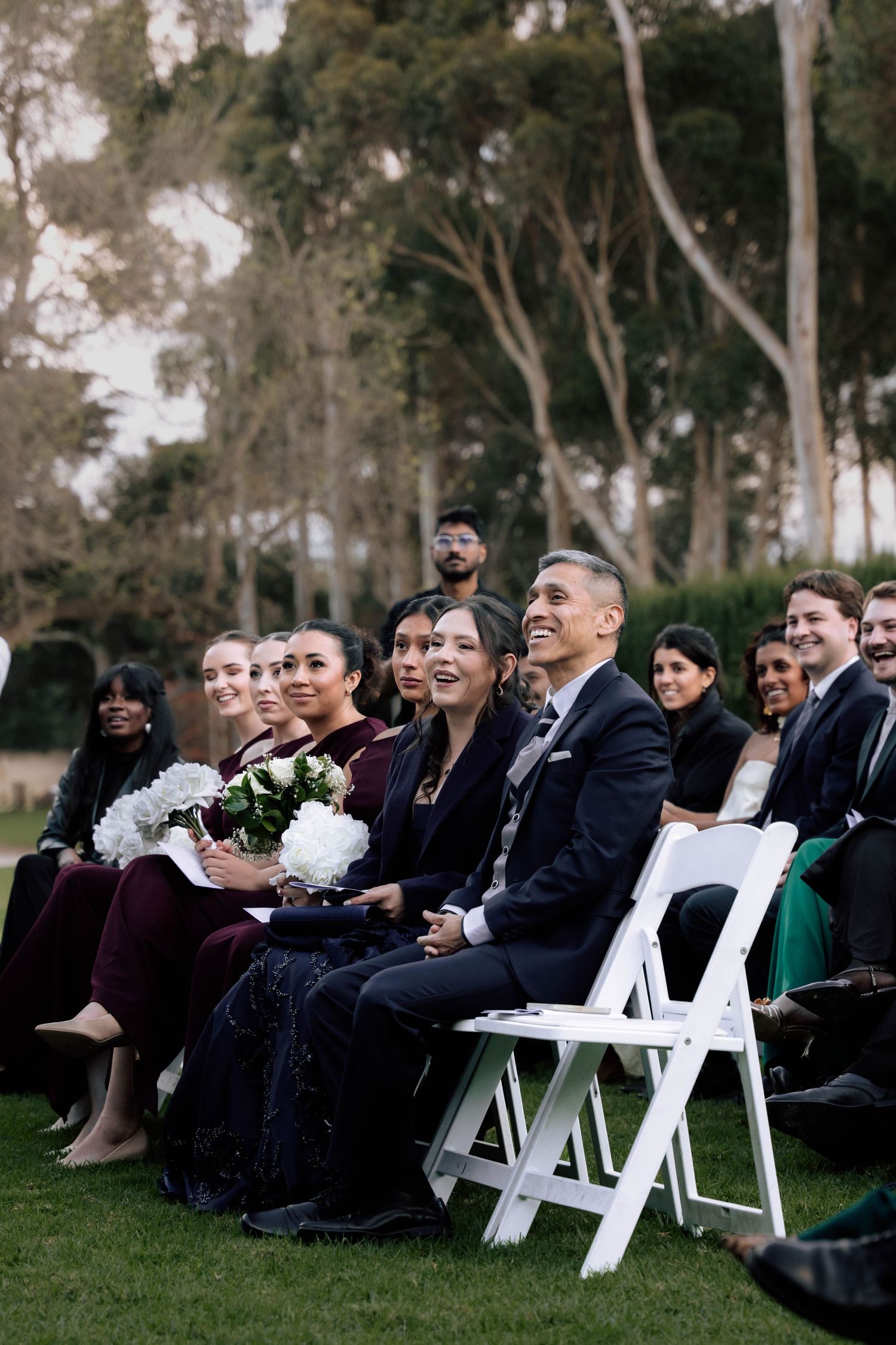 Wedding guests seated on white chairs outdoors smile and laugh during ceremony on lush green lawn.