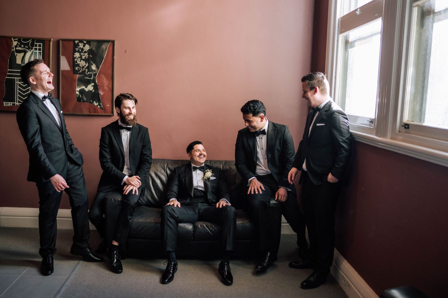 Groom and groomsmen in black tuxedos laughing together on a leather couch before the wedding ceremony.