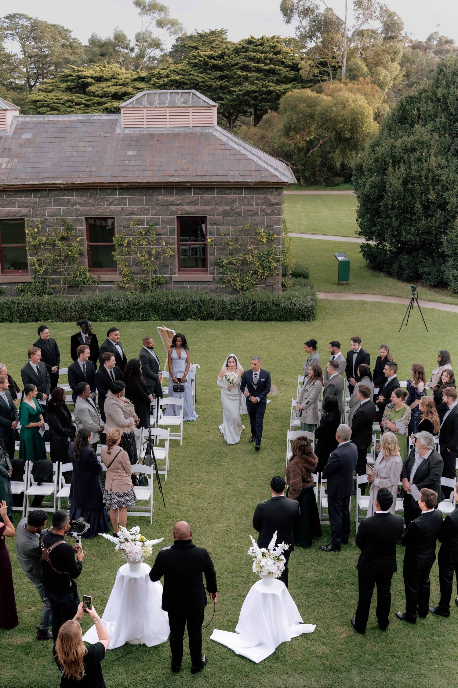Aerial view of outdoor wedding ceremony on green lawn beside historic stone building.