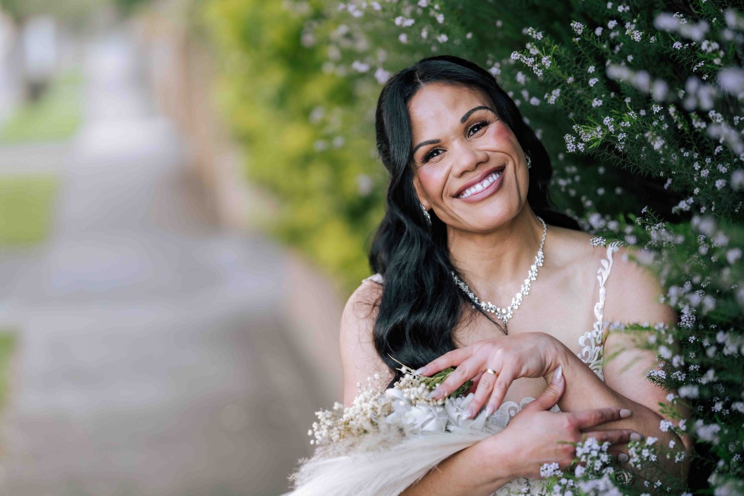 Smiling bride with dark hair holding a white bouquet among blooming flowers in an outdoor setting.