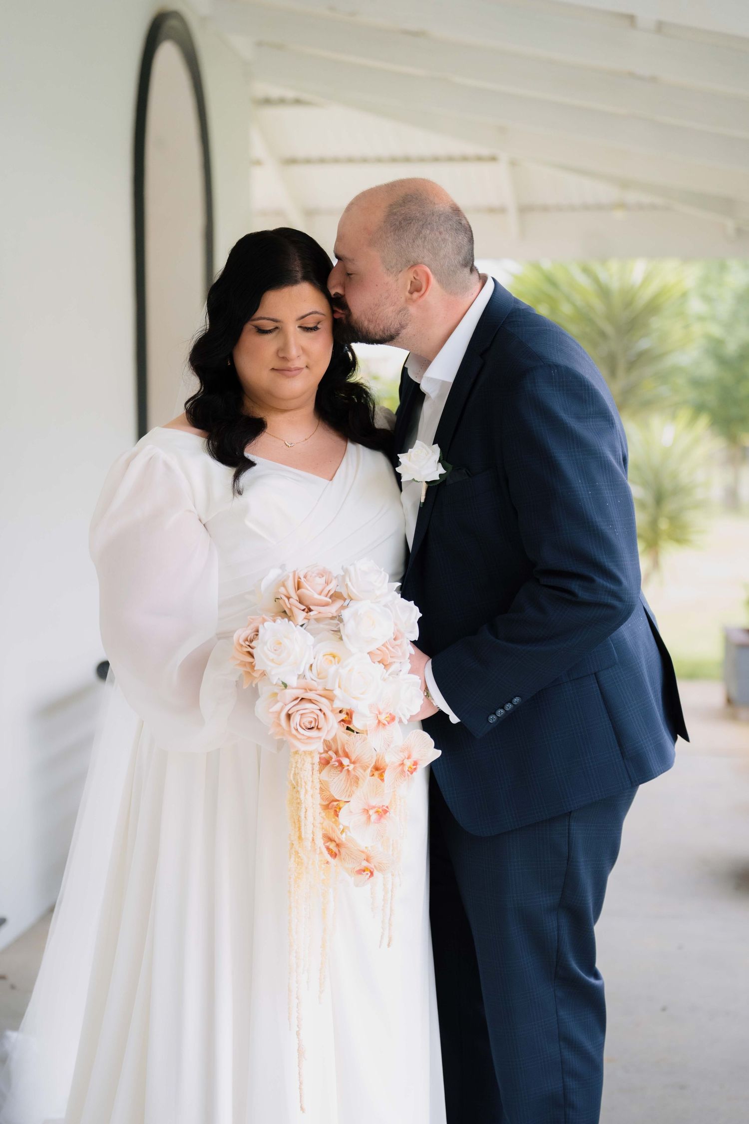 Groom in navy suit kissing bride on cheek as she holds elegant cascading peach and white rose wedding bouquet.