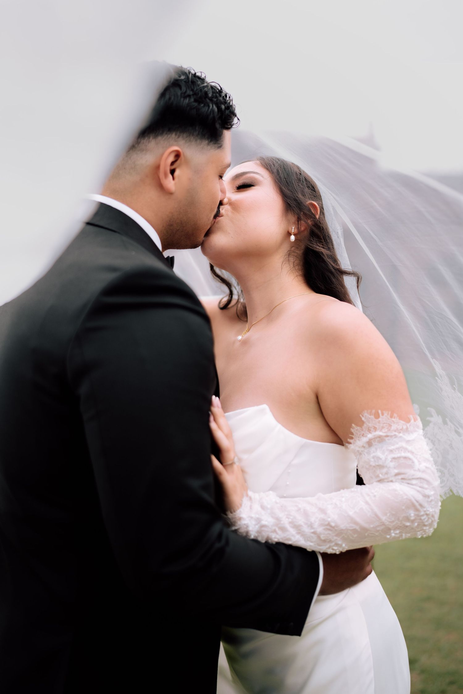 Bride and groom share romantic kiss under flowing veil in dreamy outdoor wedding portrait.