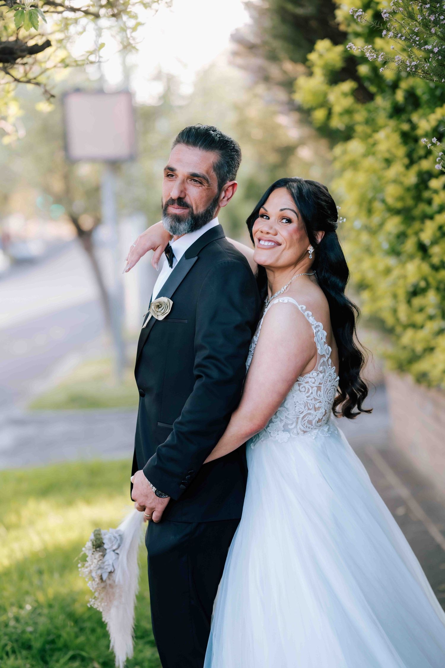 Bride and groom posing back-to-back on tree-lined street in wedding attire.