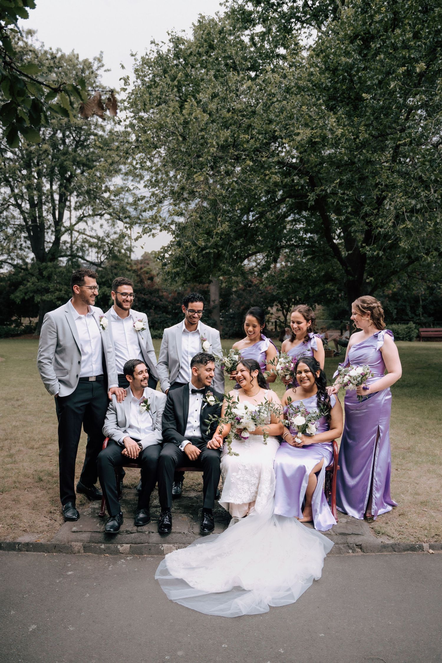 Wedding party poses in park, bridesmaids in lavender gowns and groomsmen in gray suits with couple.