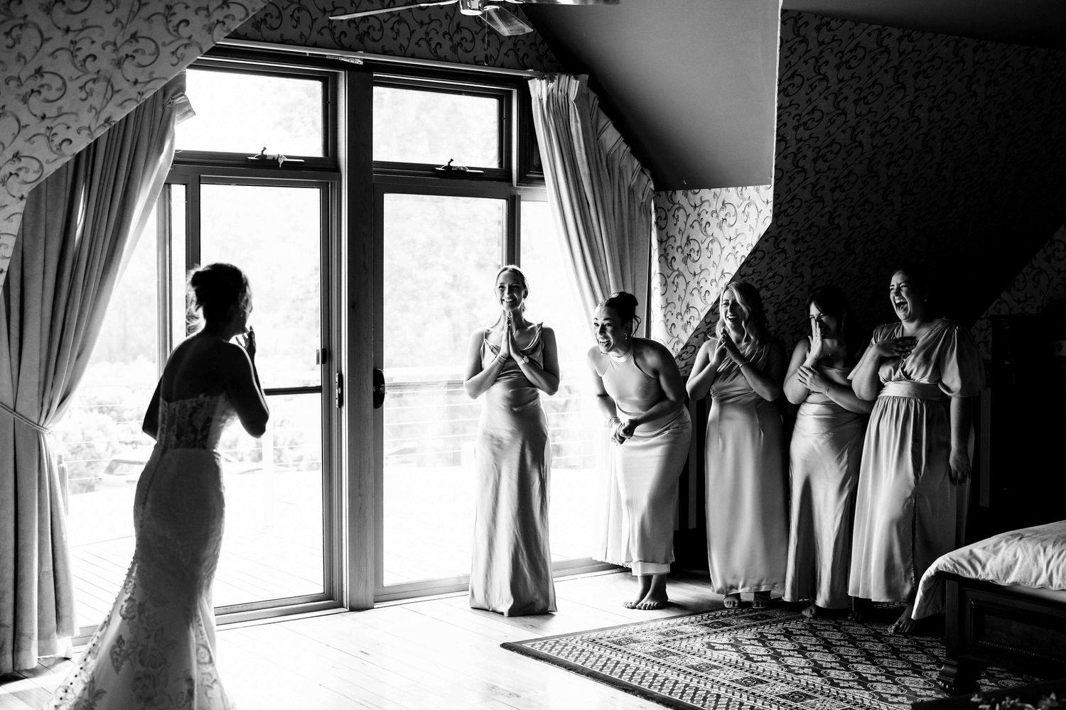 Black and white photo of bridesmaids reacting with joy as bride reveals wedding dress.