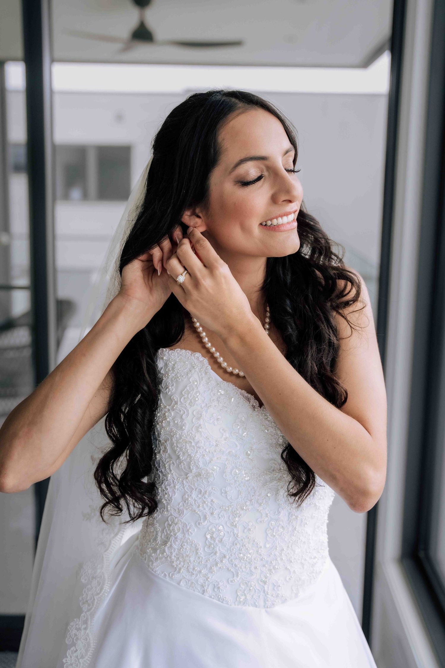 Bride smiling with eyes closed, adjusting pearl earring, wearing white lace wedding gown and pearl necklace.