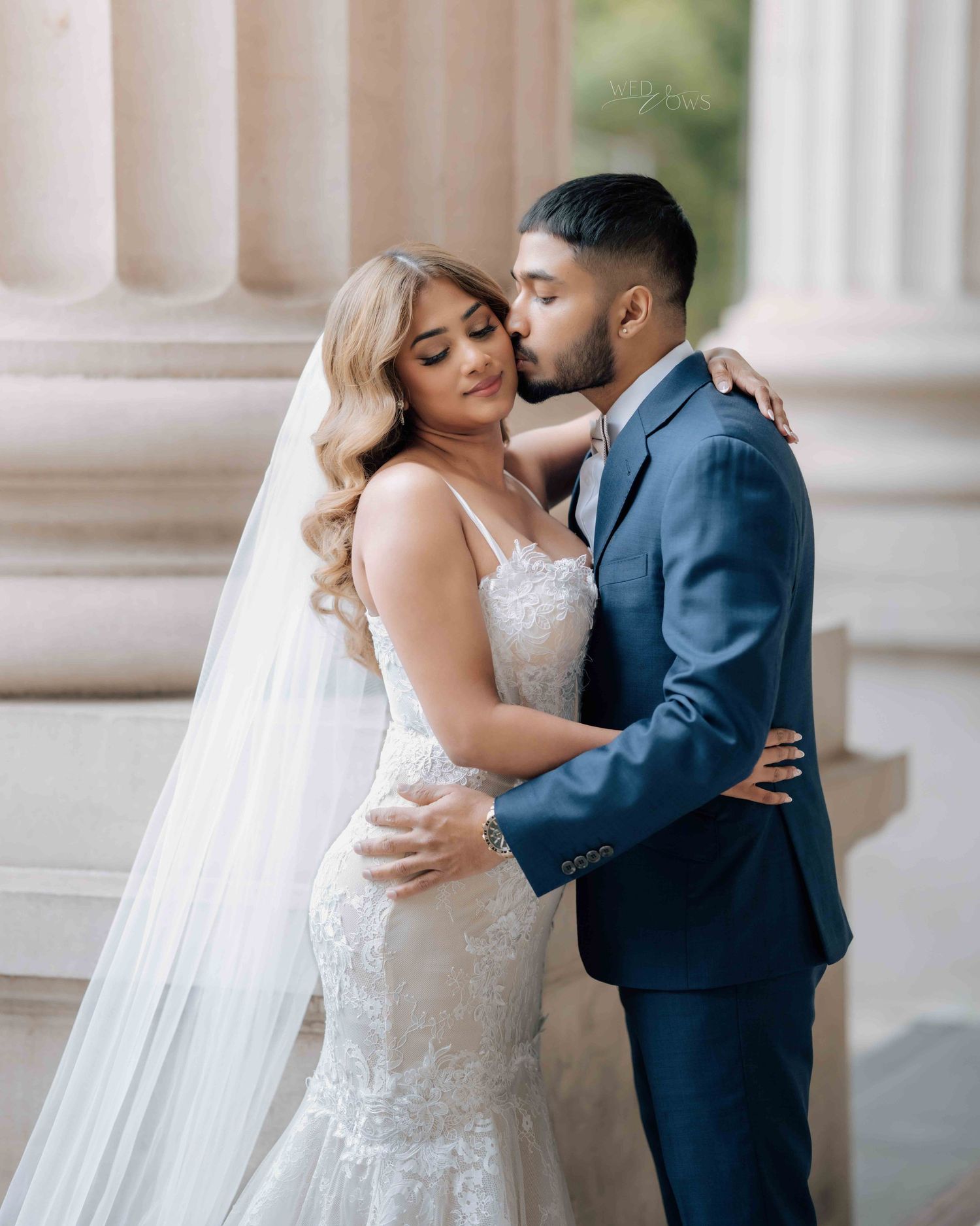 Bride in lace mermaid gown with veil as groom in navy suit kisses her forehead by white columns.