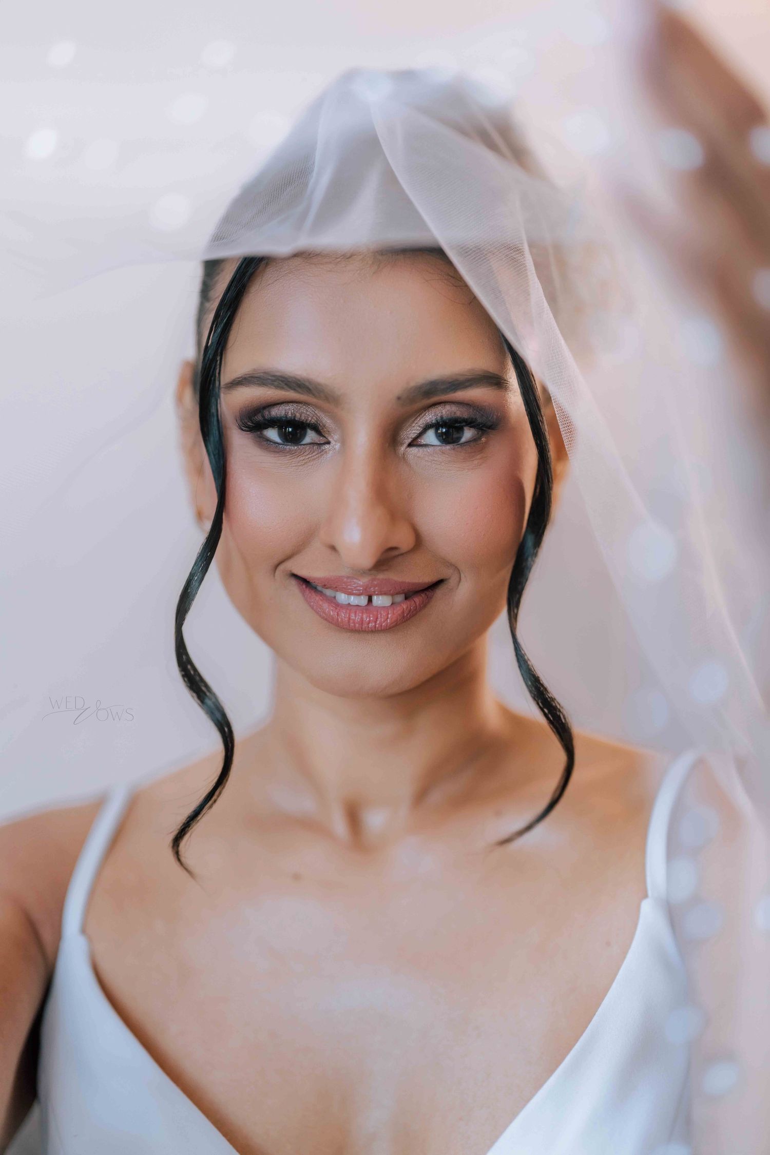 Beautiful bride smiling under a sheer white veil with glamorous makeup and dark loose tendrils.