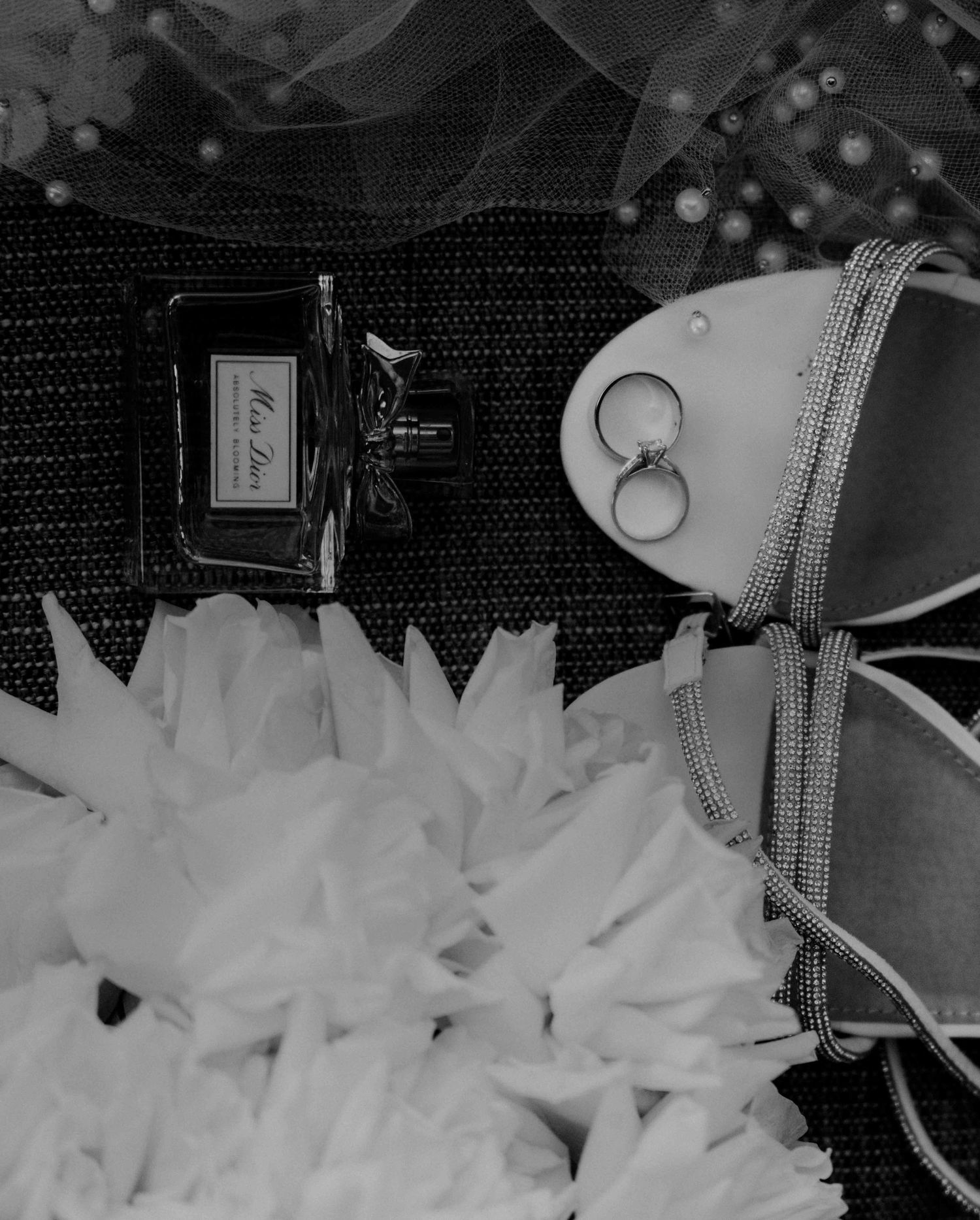 Black and white flat lay of wedding rings, bridal heels, perfume bottle and white floral bouquet.