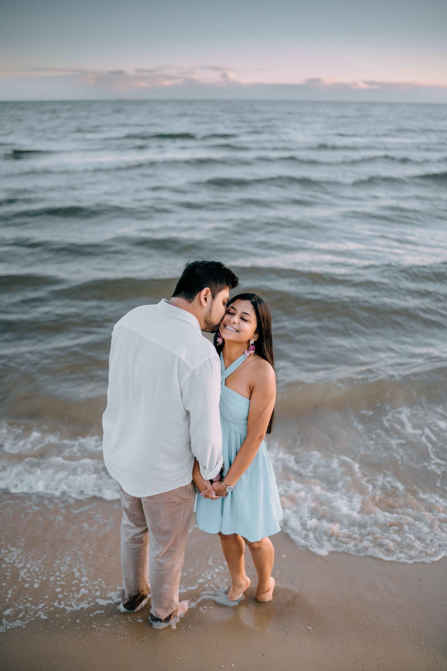 Romantic couple holding hands at the beach shoreline at sunset, man kissing woman in light blue dress.