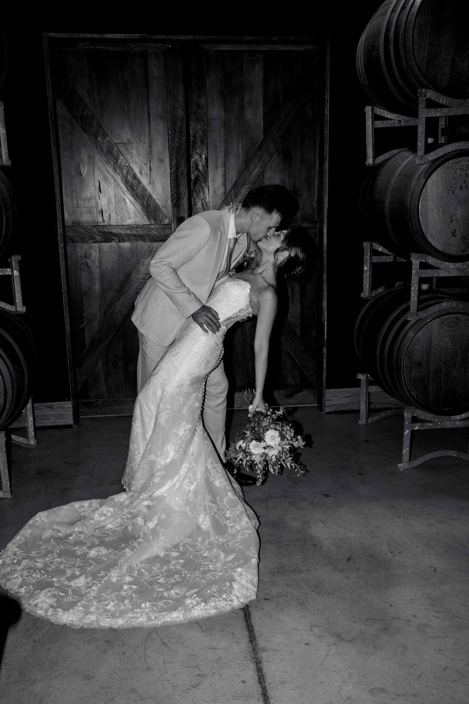 Groom dips bride for romantic kiss near wine barrels in black and white winery wedding portrait.