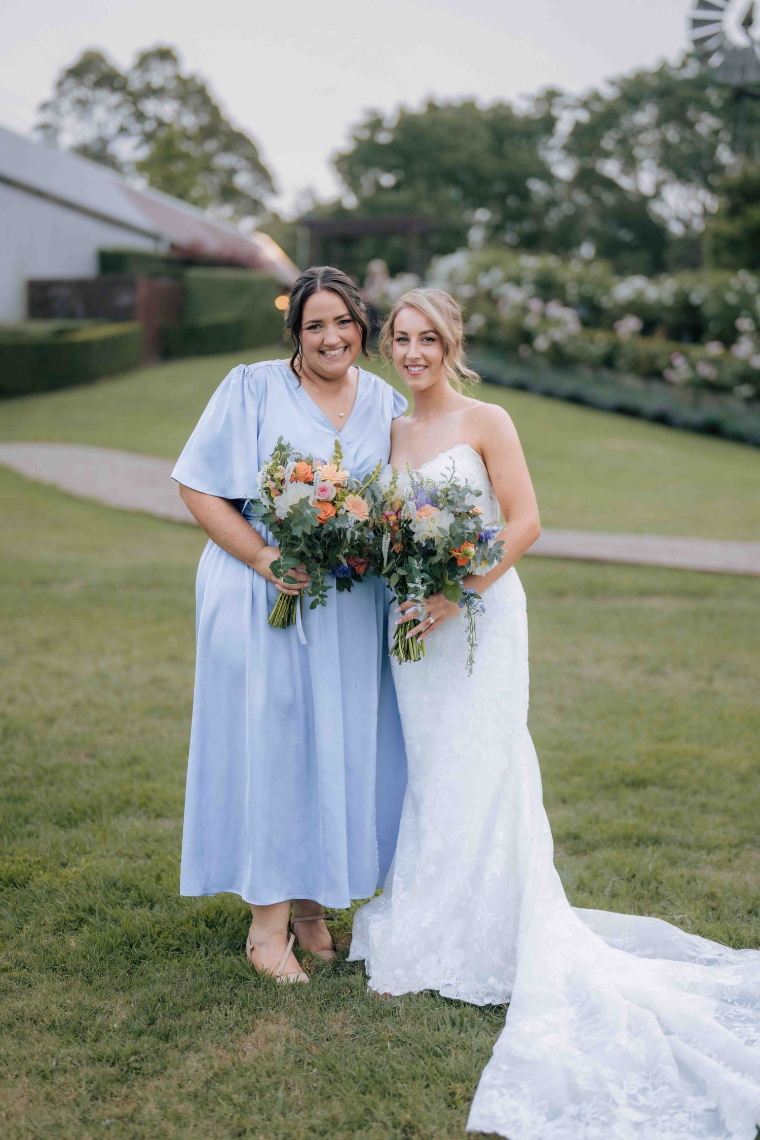 Bride in white lace mermaid gown poses with bridesmaid in light blue midi dress holding colorful floral bouquets.