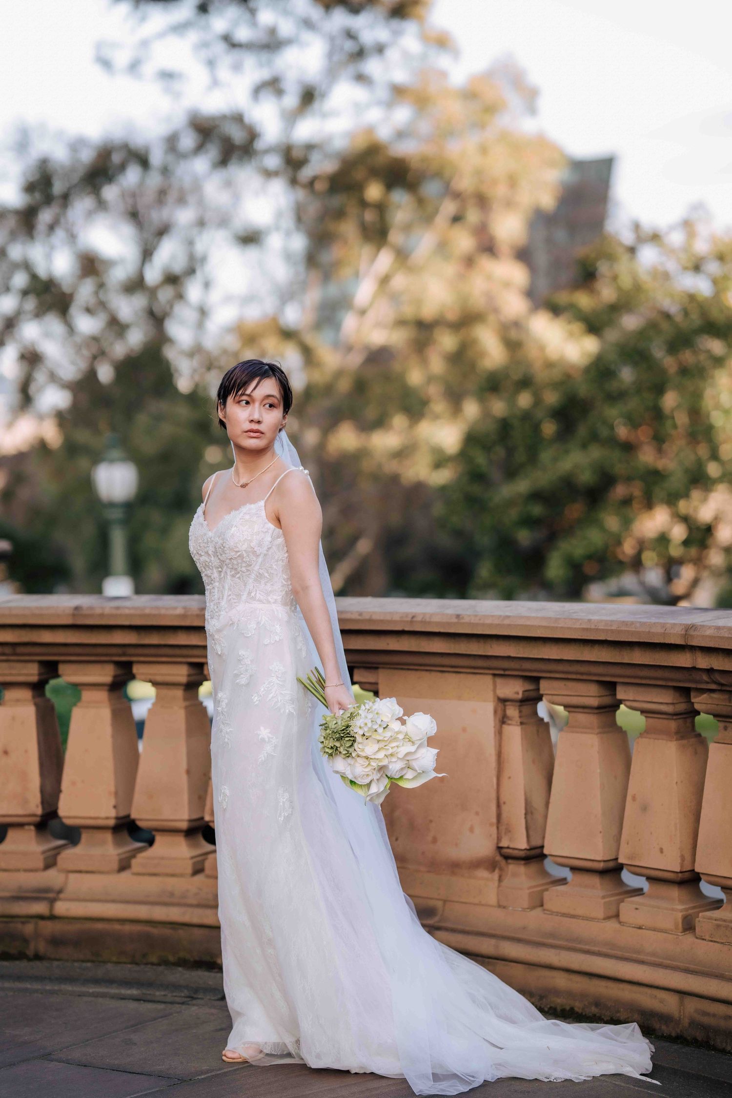 Bride in white lace wedding gown with veil holds bouquet by stone balustrade in park.