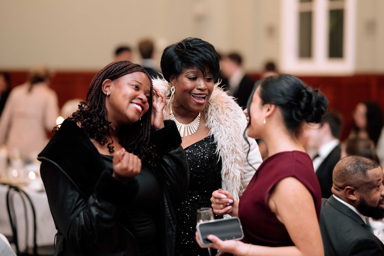Three women laughing and embracing joyfully at elegant wedding reception dinner.