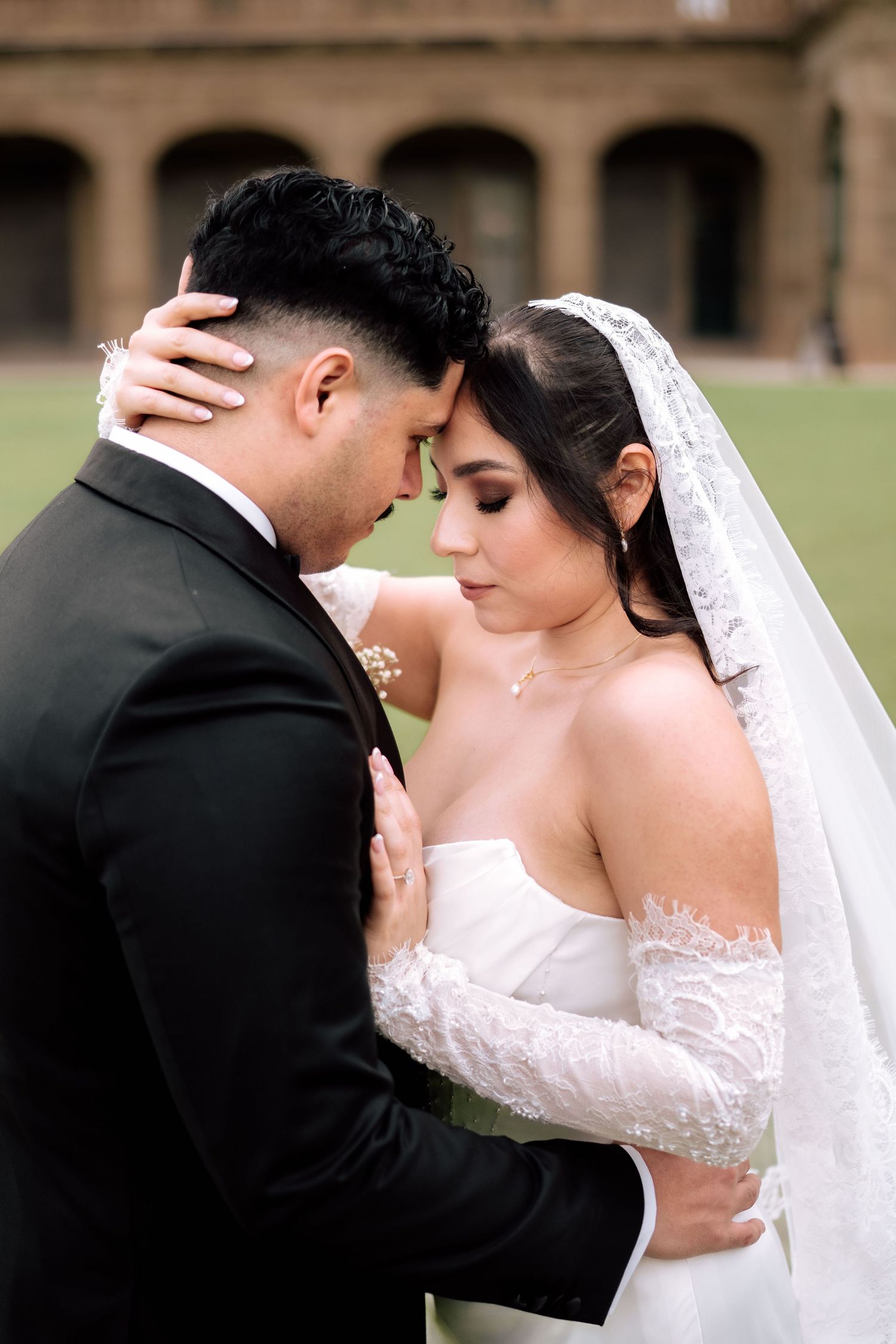 Bride in lace gown and groom in black tuxedo share intimate moment outdoors.