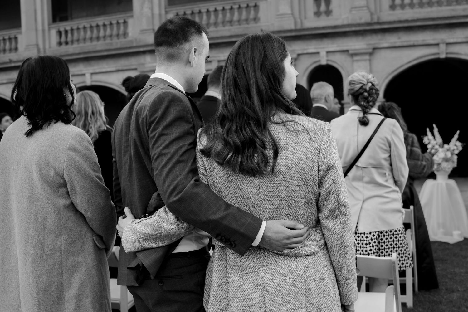 Black and white photo of couple embracing while standing among wedding guests at outdoor ceremony.