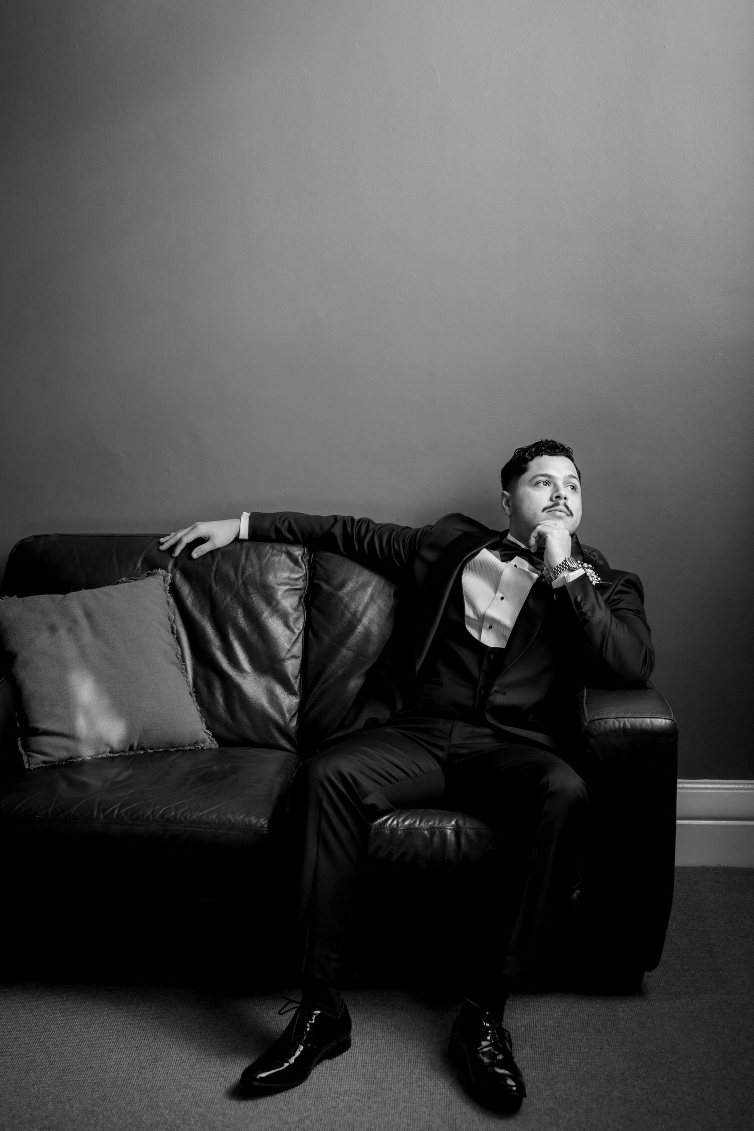 Elegant man in tuxedo sitting pensively on leather sofa in black and white portrait photo.
