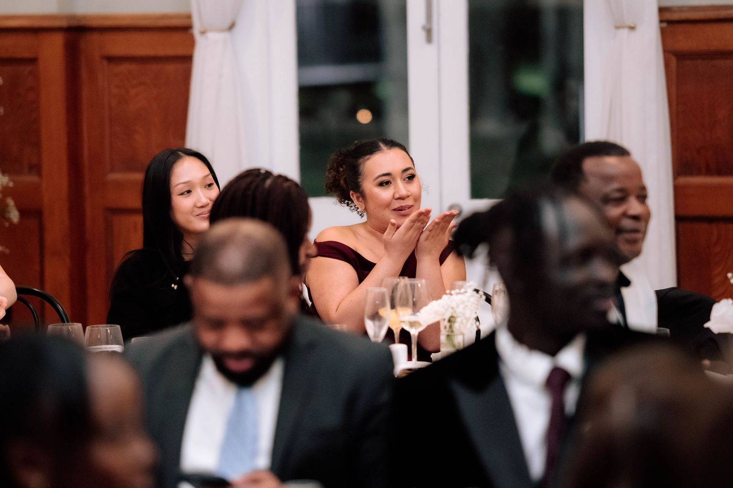 Wedding guest in burgundy dress clapping and smiling during a speech at an elegant reception.