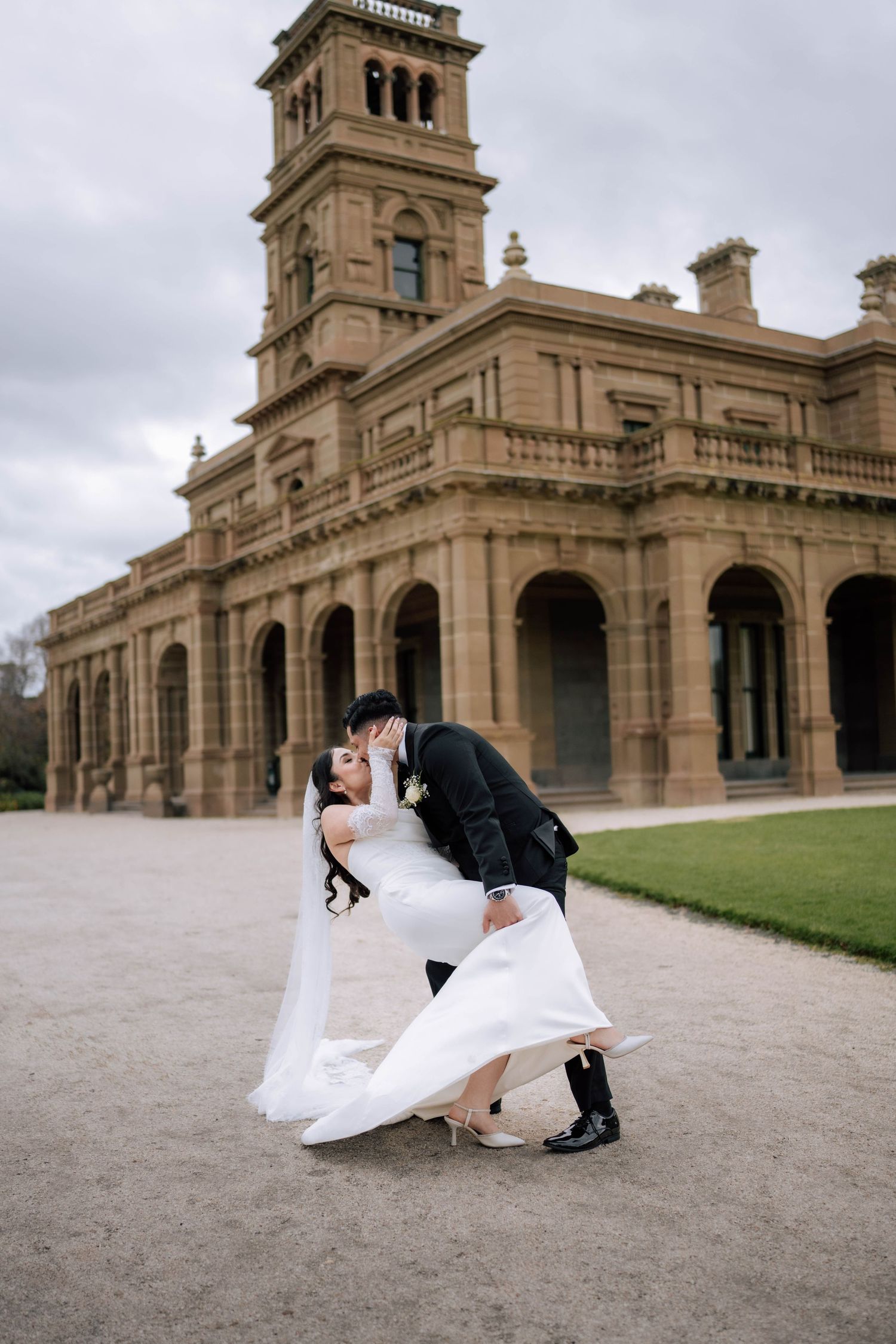 Bride and groom share a romantic dip kiss in front of a grand historic stone mansion on their wedding day.