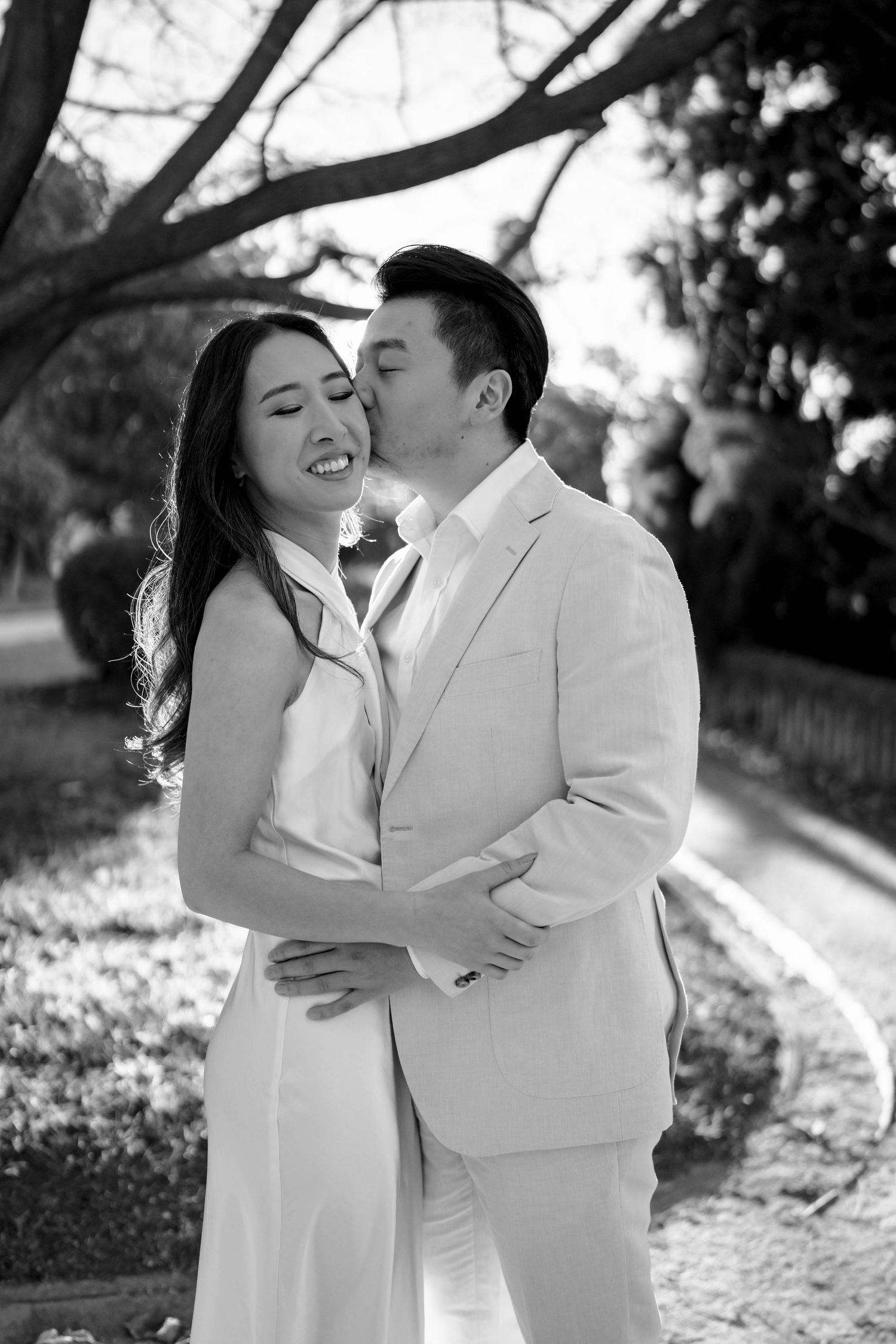 Black and white photo of Asian groom kissing bride on cheek while embracing in a sunlit park setting.