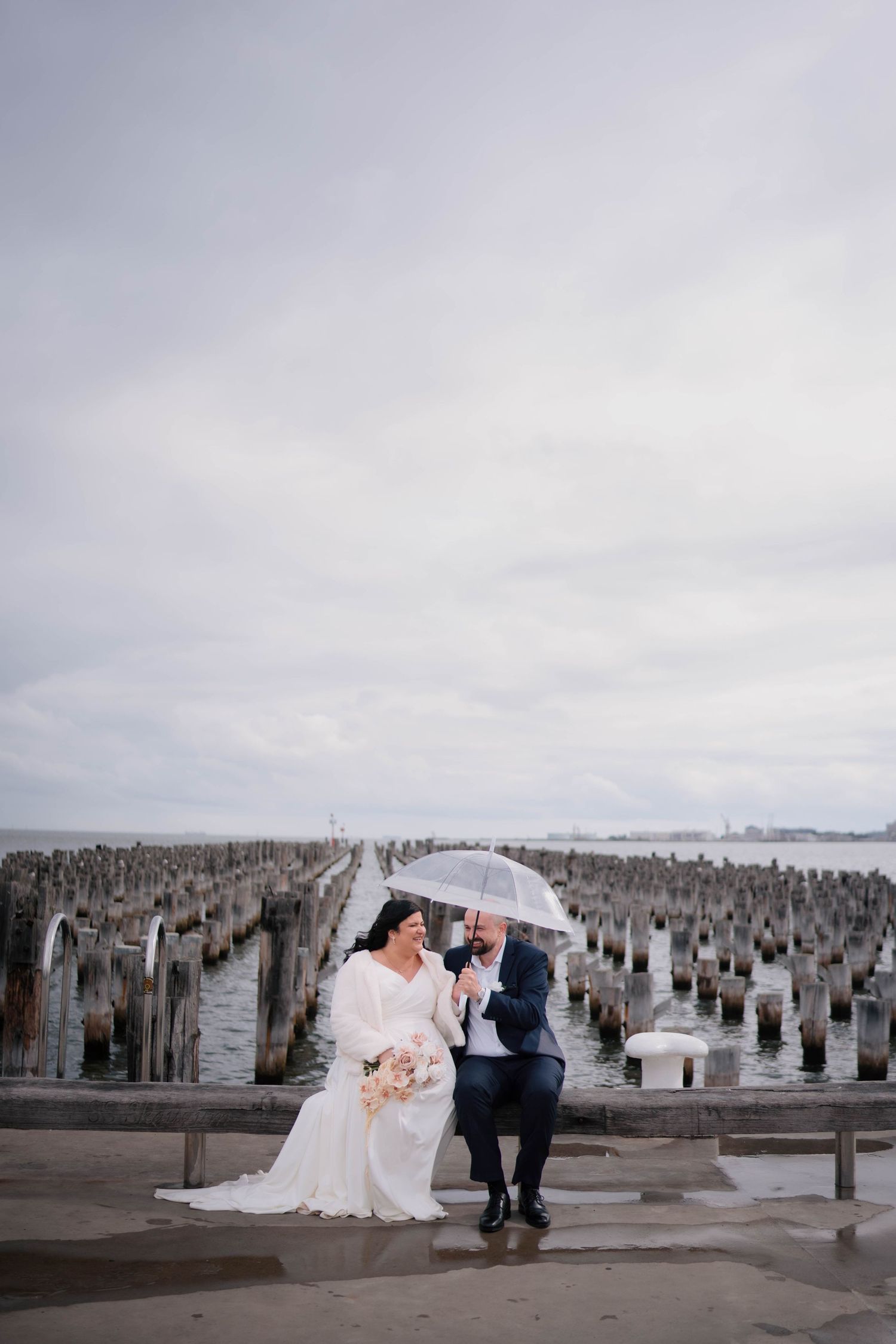 Bride and groom sit on a waterfront pier bench sharing a clear umbrella on a cloudy wedding day.