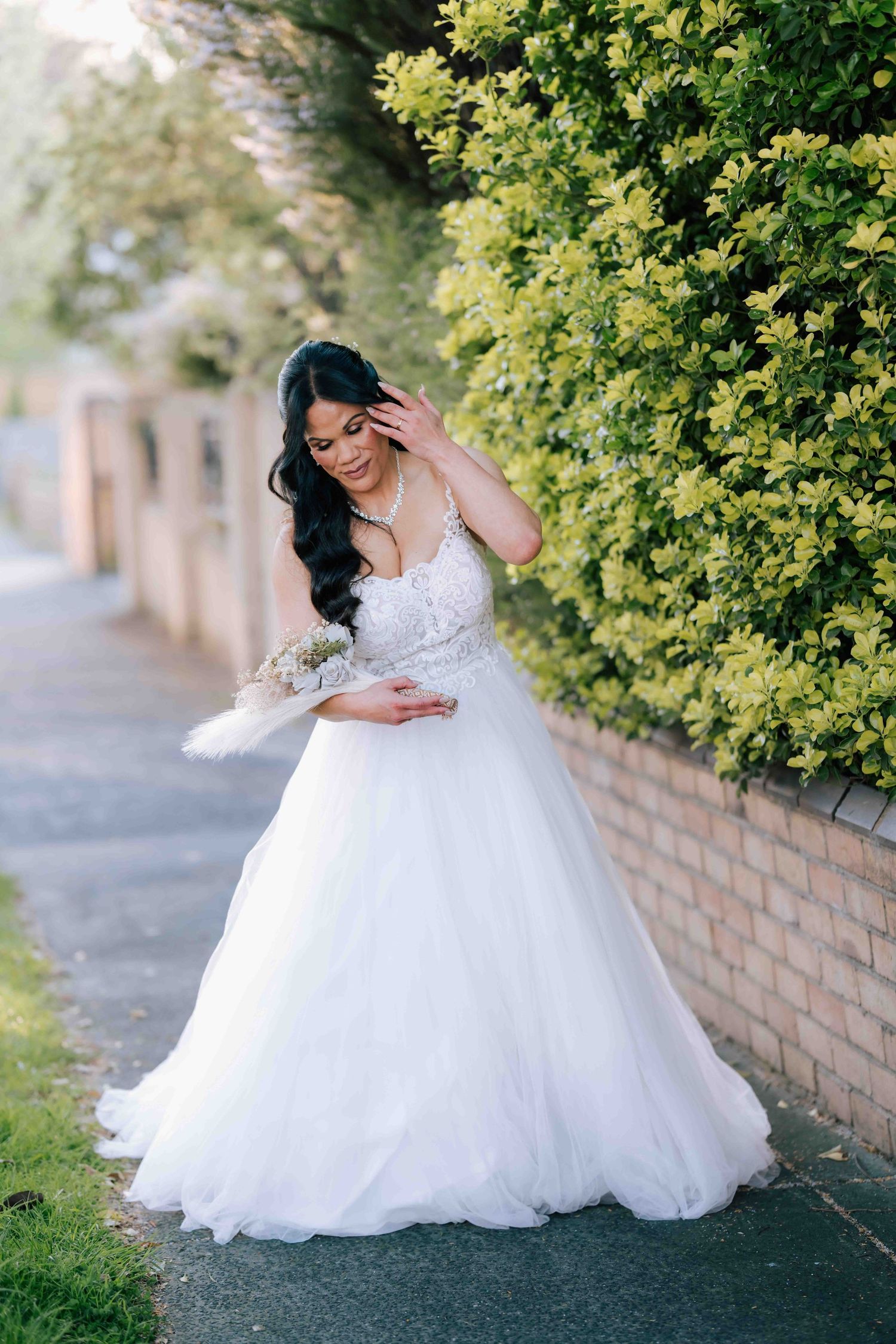 Elegant bride in white ballgown standing beside lush green hedge on sunny day.