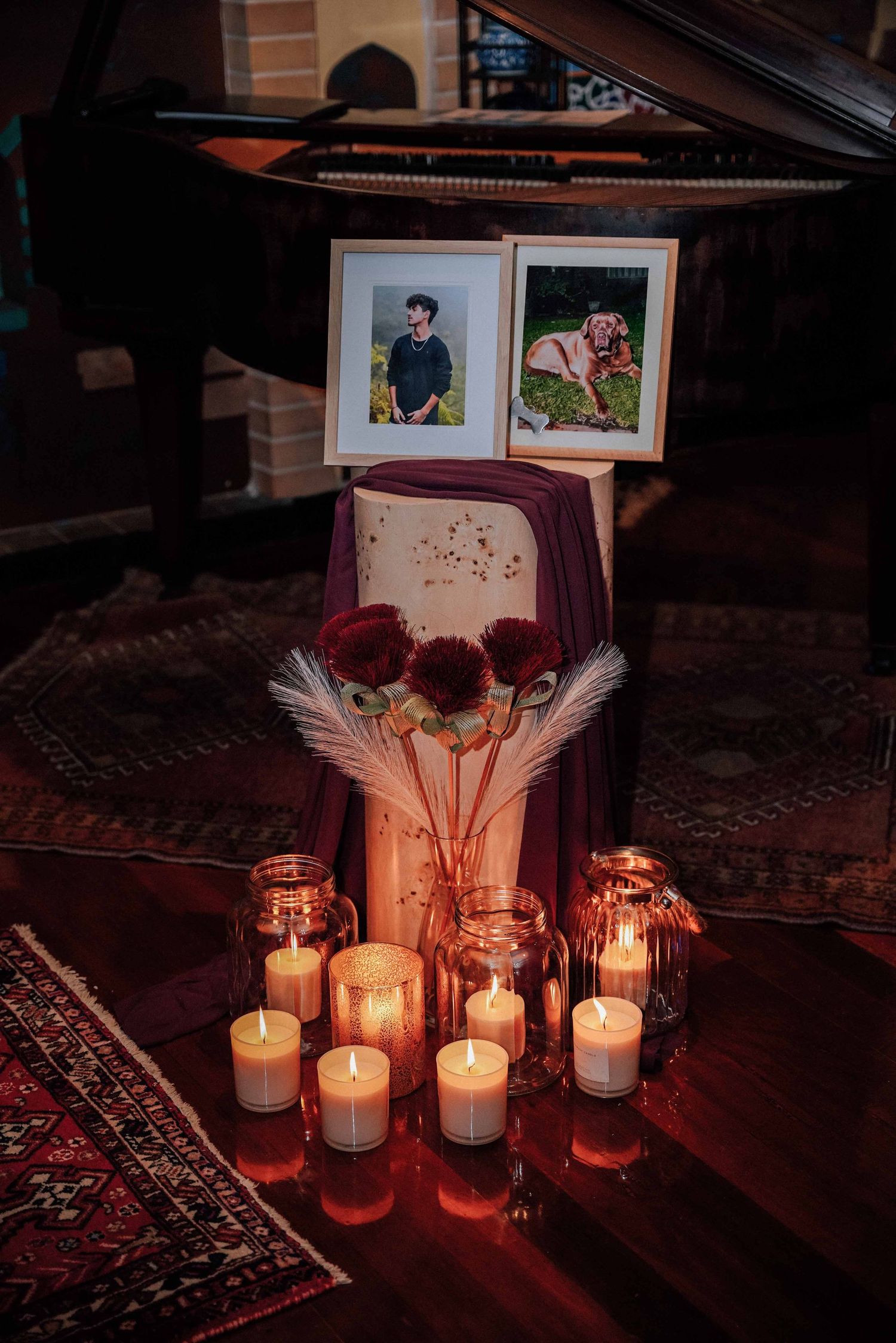 Wedding memorial display with framed photos, red florals, pampas grass, and glowing candles near piano.