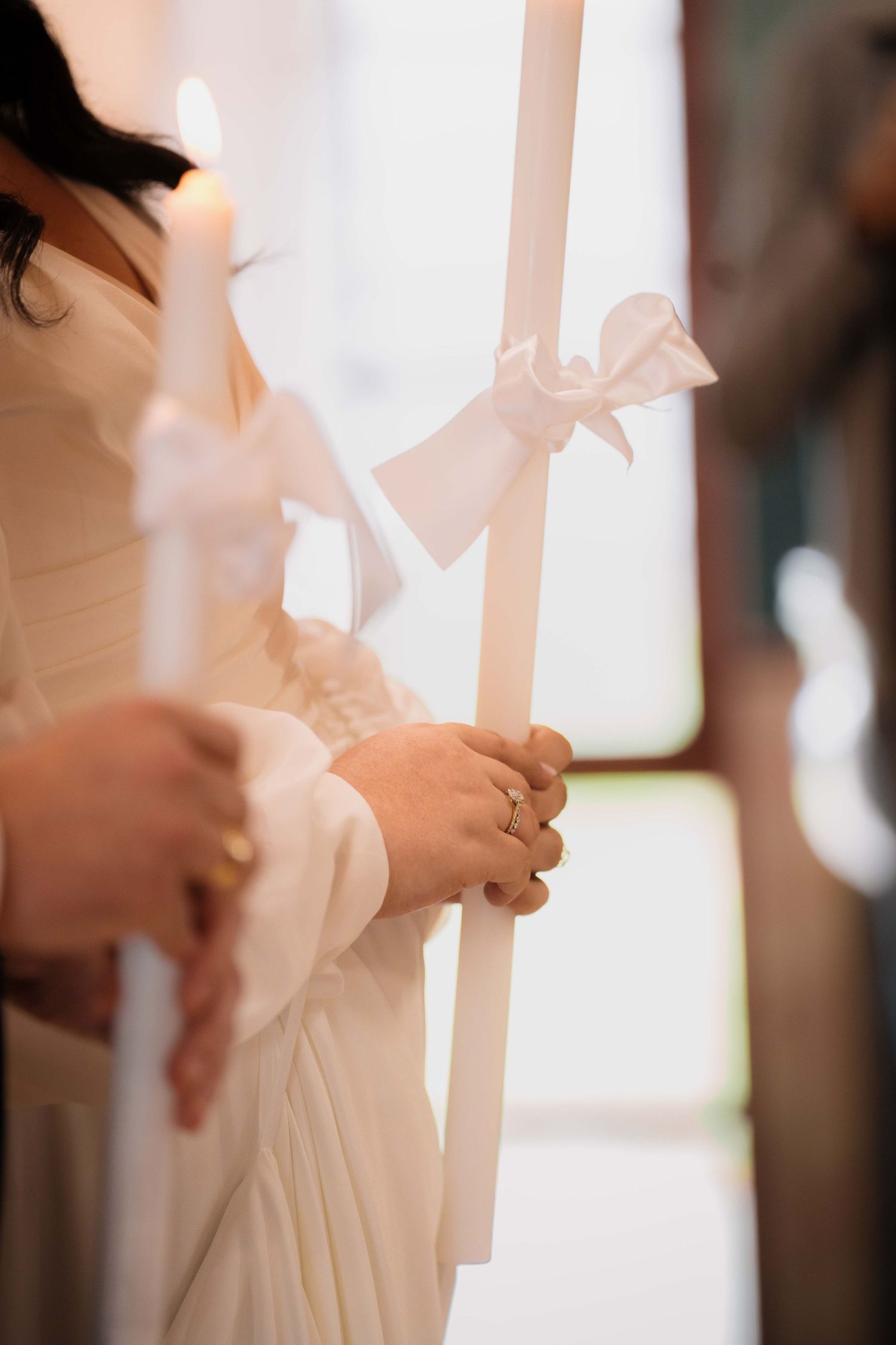 Bride in white dress holding tall white wedding candles adorned with satin ribbons during a church ceremony.