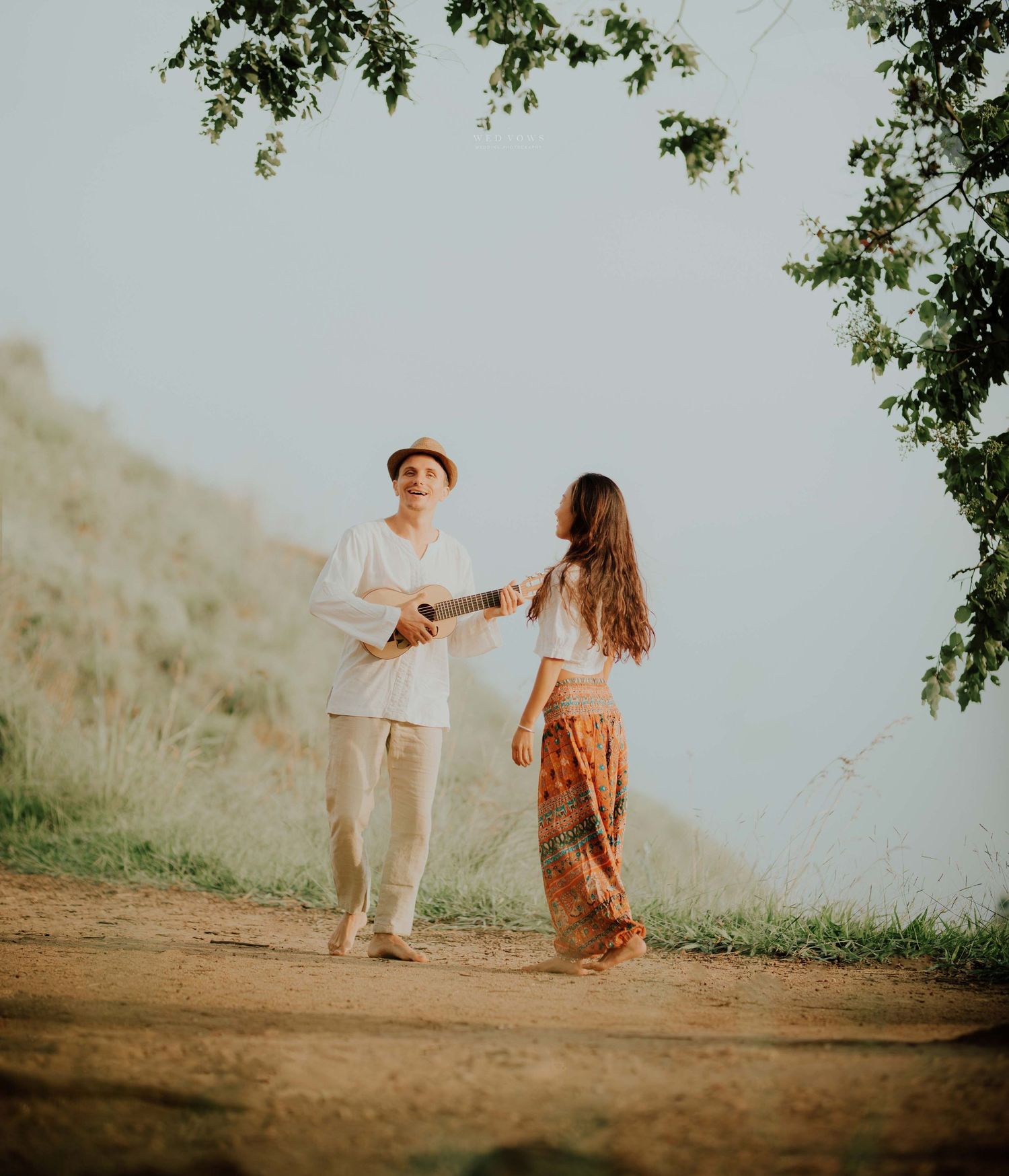 A couple holding hands on a scenic dirt path, man wearing a hat and white outfit, woman in bohemian skirt.